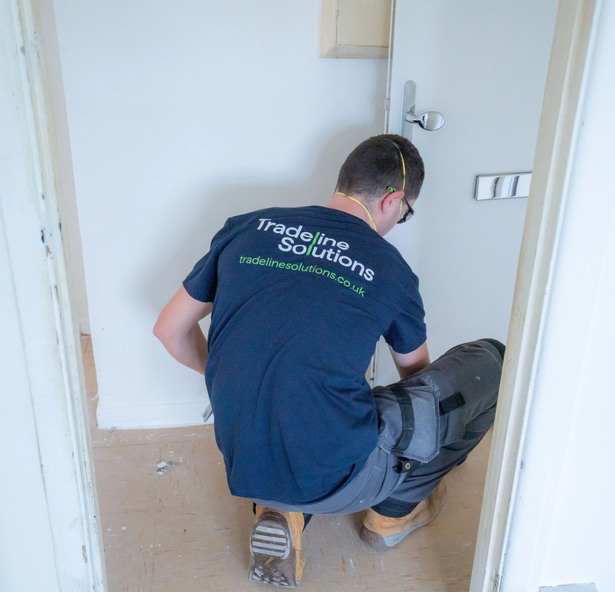 A man working on a small construction project inside a doorway, wearing a blue T-shirt with green and white text on the back, tan work boots, and glasses. The room has light-colored walls and a wooden floor with some debris and tools on it.