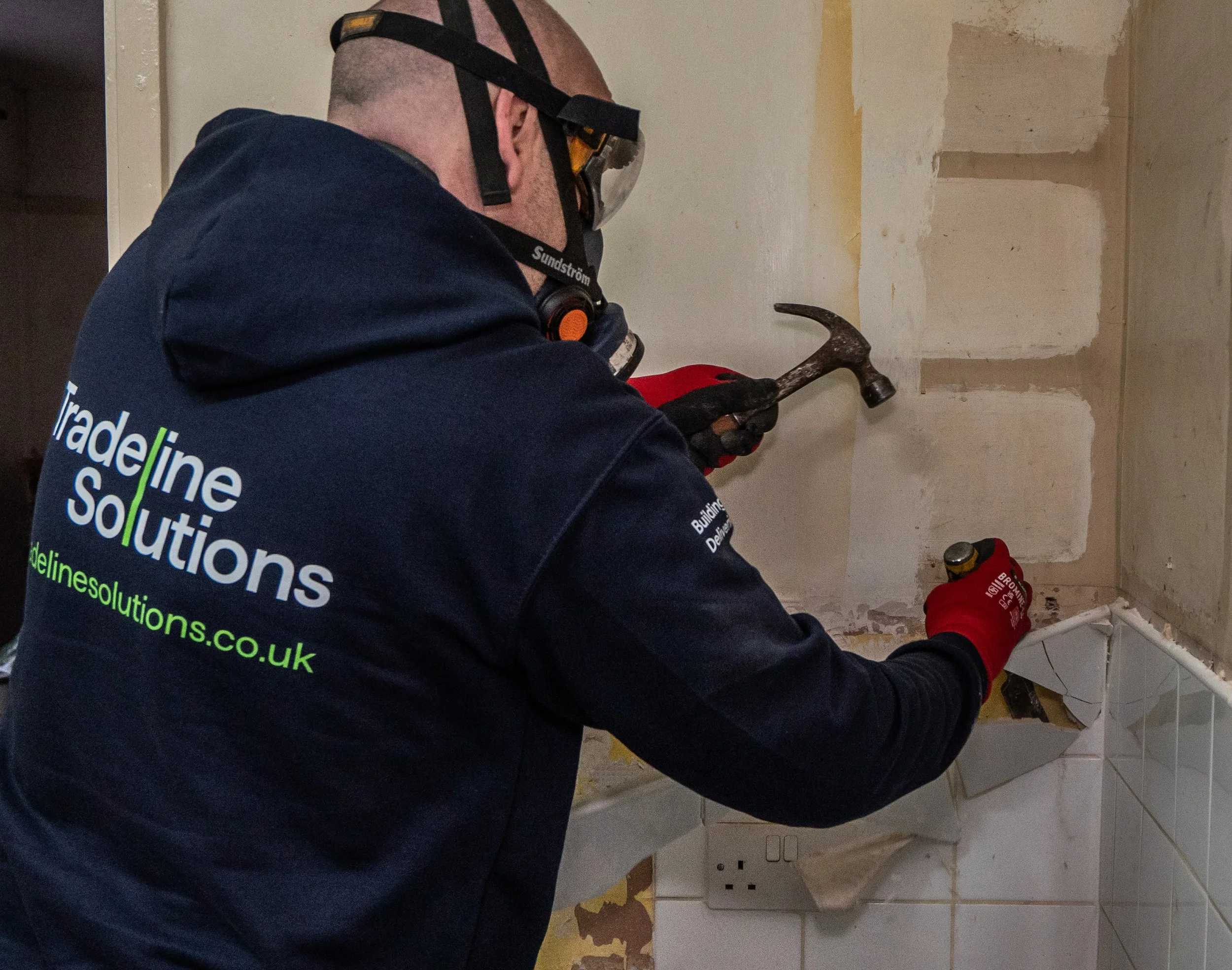 A construction worker wearing safety goggles and red gloves is using a hammer to remove or break tiles from a wall in a room under renovation.