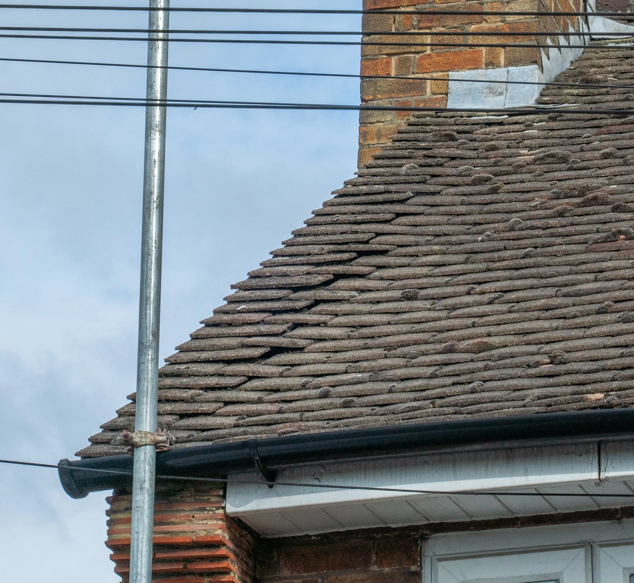 Close-up of a rooftop with reddish-brown clay tiles under a cloudy sky, featuring a chimney made of bricks, a metal scaffolding pole, and wires.