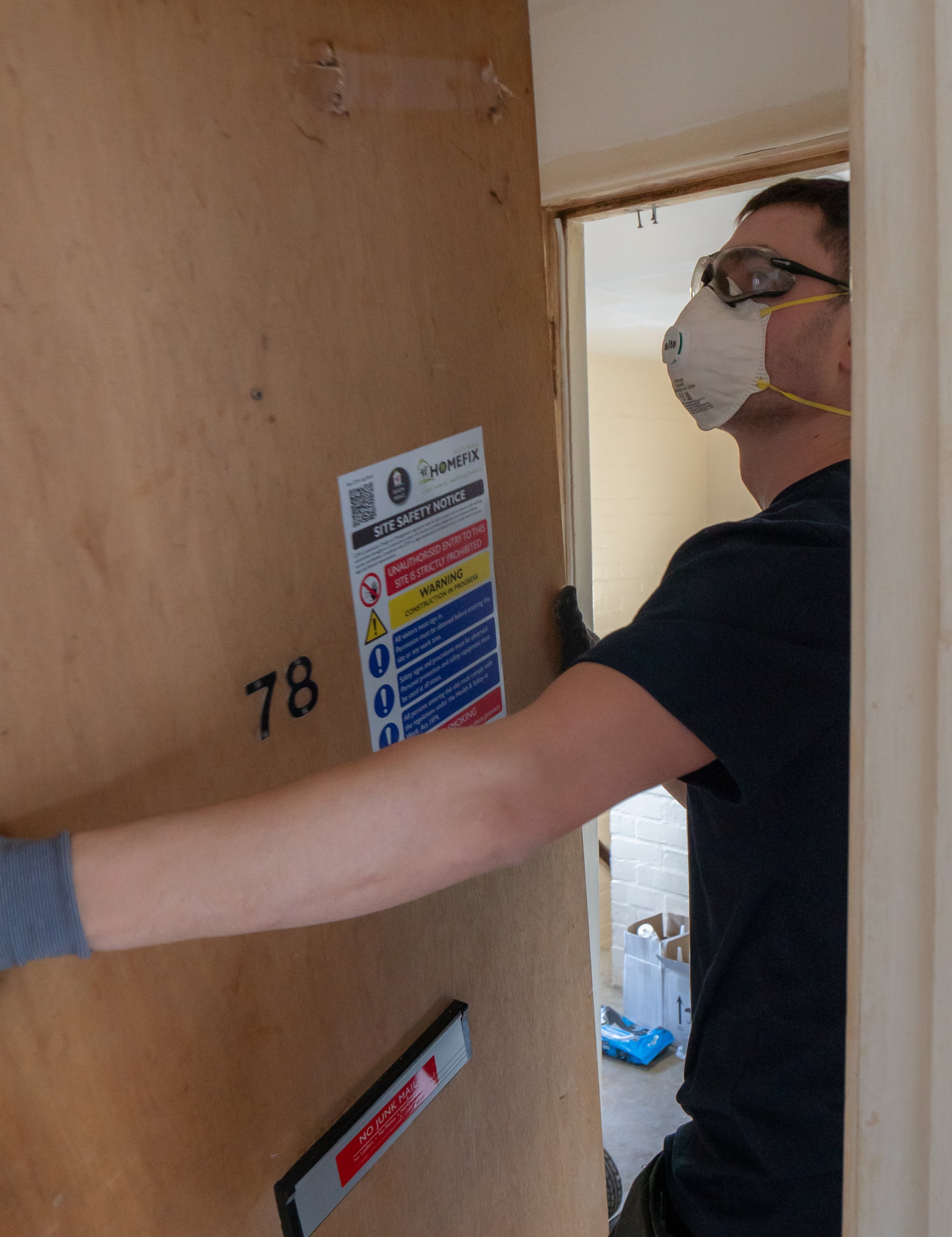 A person wearing glasses, a face mask, and a black shirt is at a postal drop box, preparing to mail a package. The drop box has safety notices and a slot for mailing.