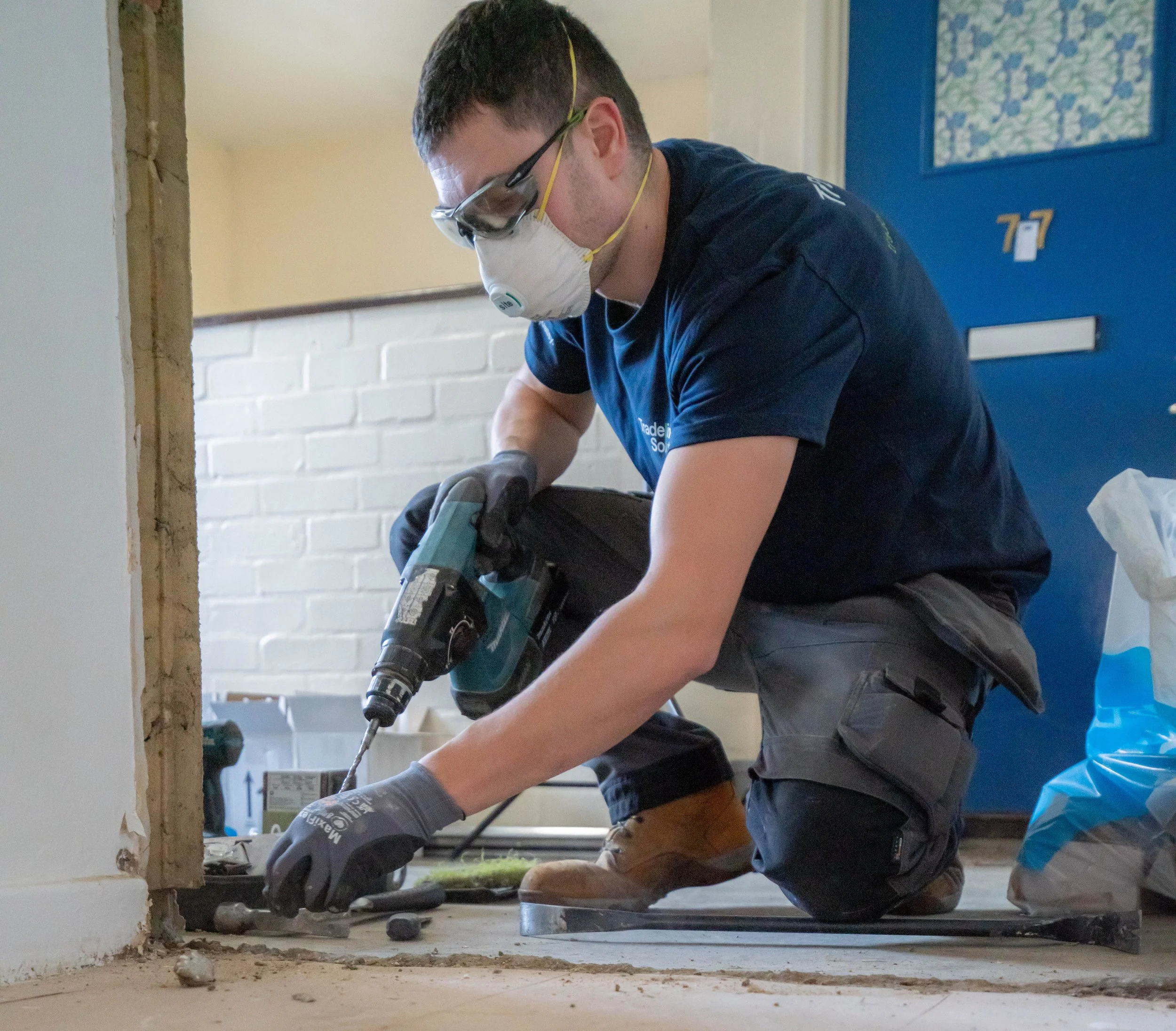 A man wearing safety goggles, a face mask, and gloves is kneeling on the floor using a power drill on a wall during a construction or renovation project.