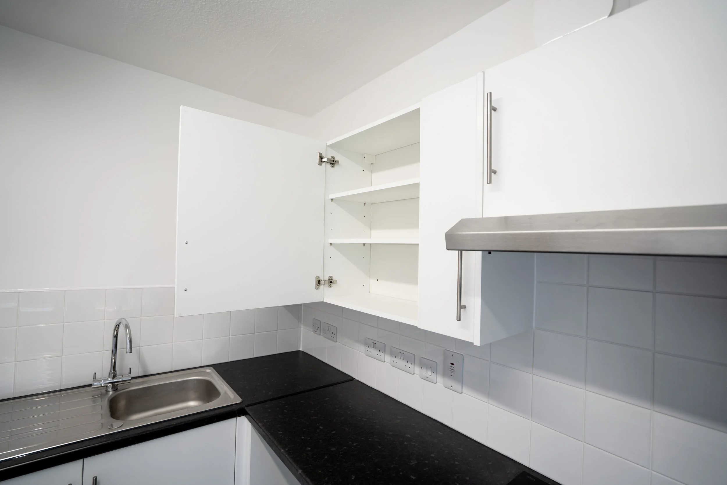 A kitchen cabinet with open doors, showing two white interior shelves, above a black granite countertop with a corner sink and a modern chrome faucet, set against a white tiled backsplash with electrical outlets.