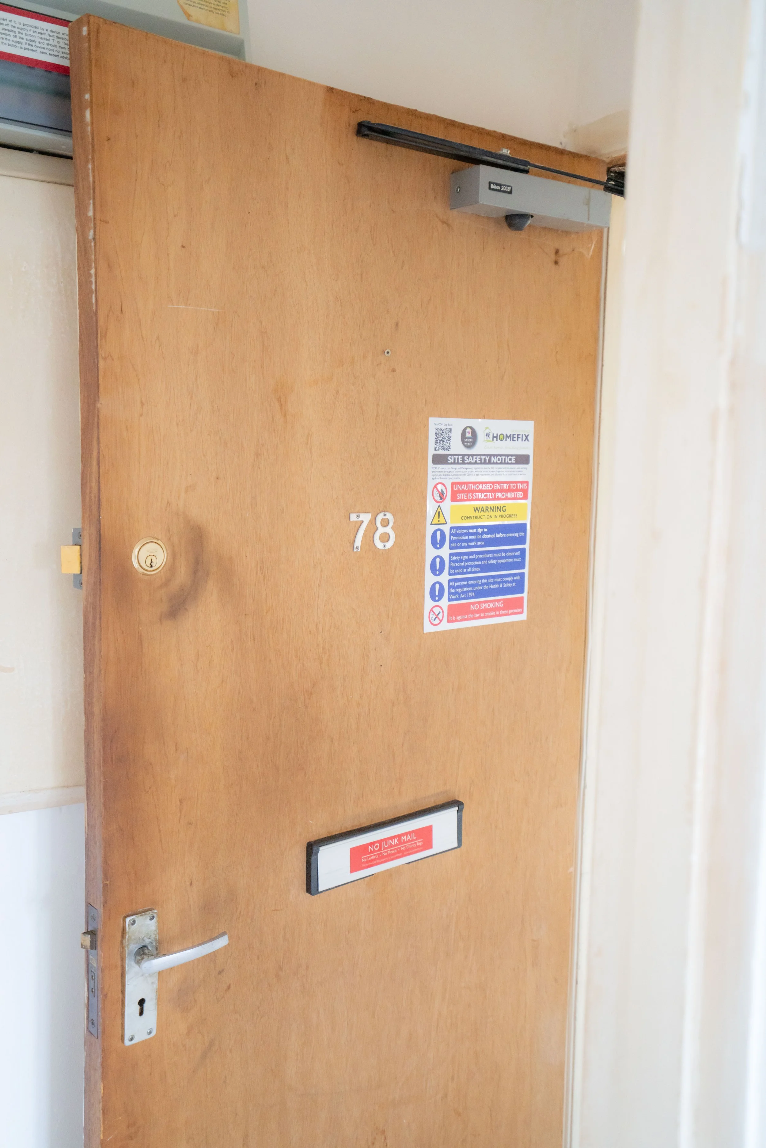 A wooden door with a silver handle and multiple safety and security stickers, including 'No Junk Mail' and 'Site Safety Notice' warnings.