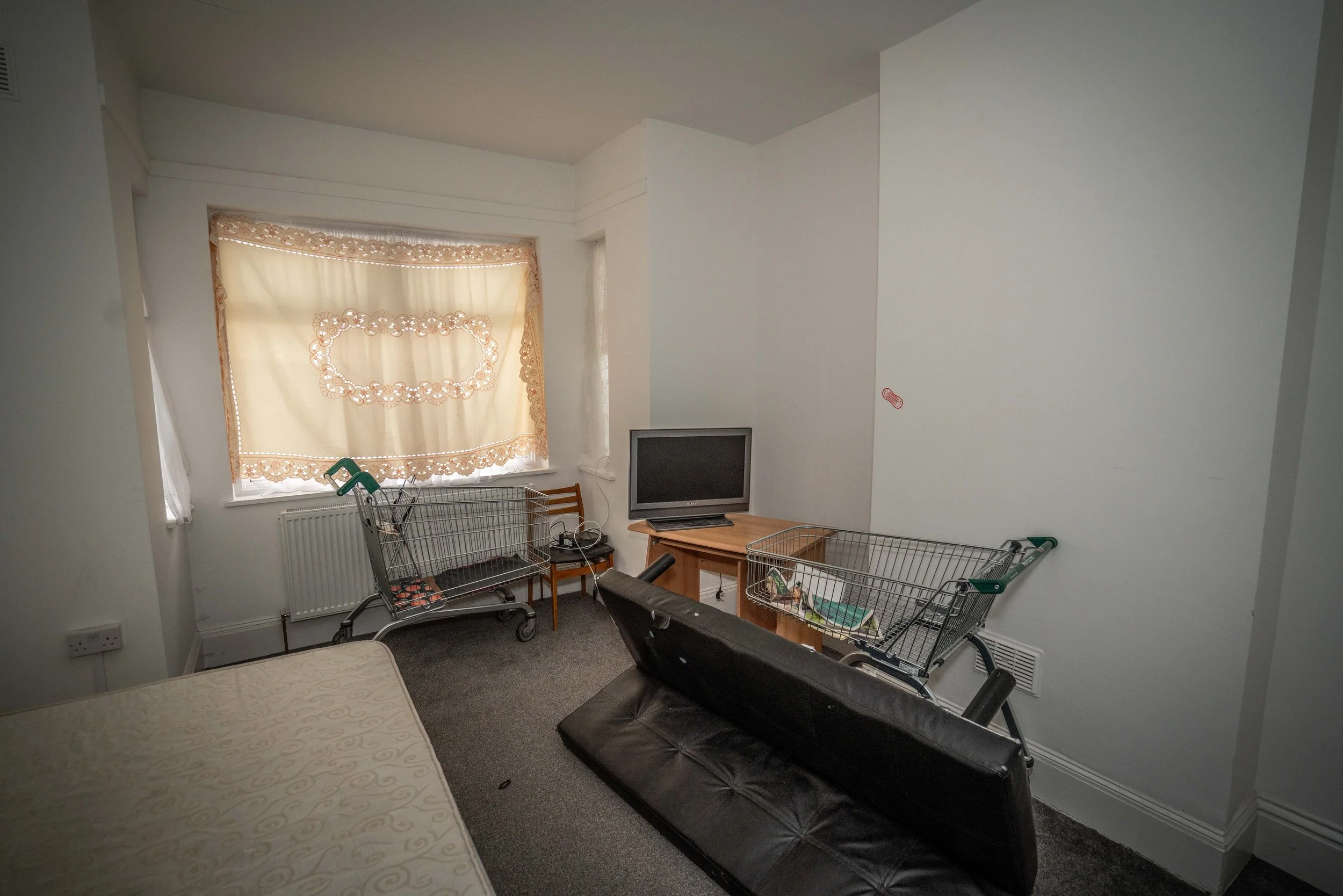 A small, cluttered living room with white walls and a window with beige lace curtains. The room contains a black leather sofa, a small wooden table with a small TV on it, two shopping carts, and a small wooden chair. The floor is carpeted and there i