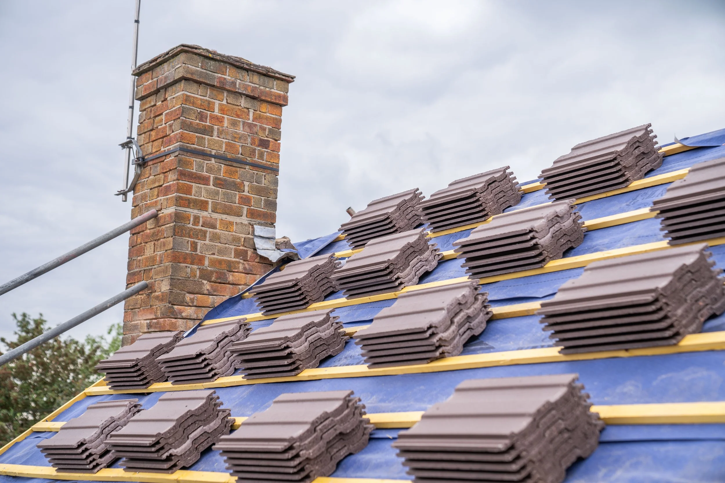 Roof under construction with brown tiles and a brick chimney against a cloudy sky.