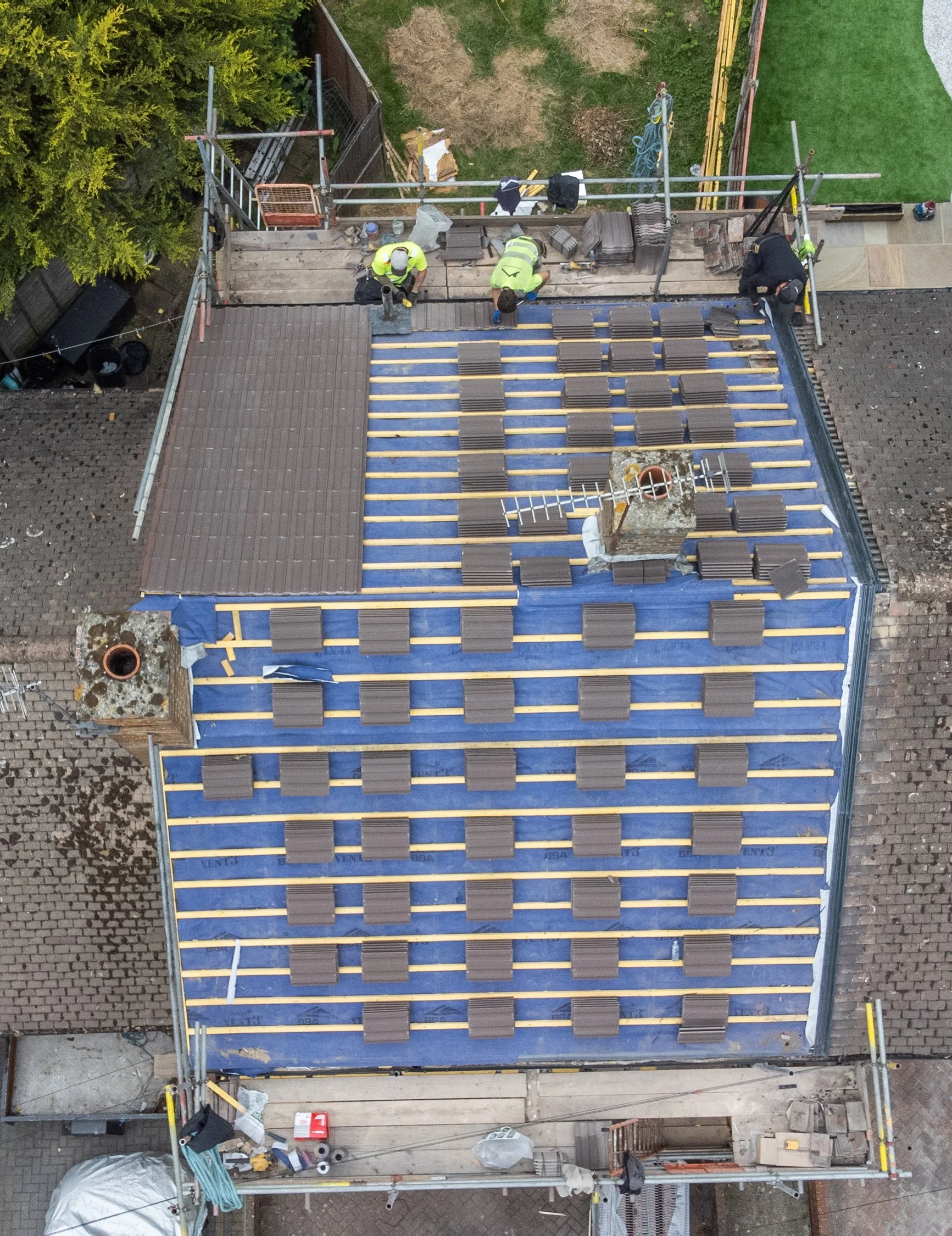 Construction workers installing tiles on a roof on scaffolding, with tools and materials around.