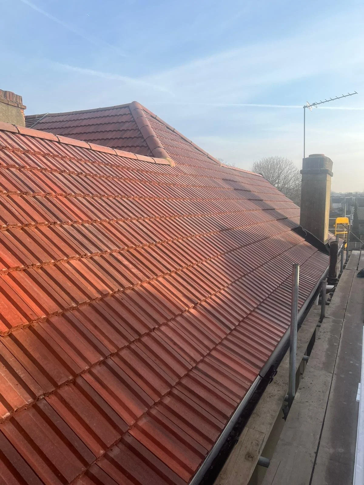 A worker wearing a yellow safety helmet, gray shirt, and reflective vest, re-caulking a roof with brown tiles.