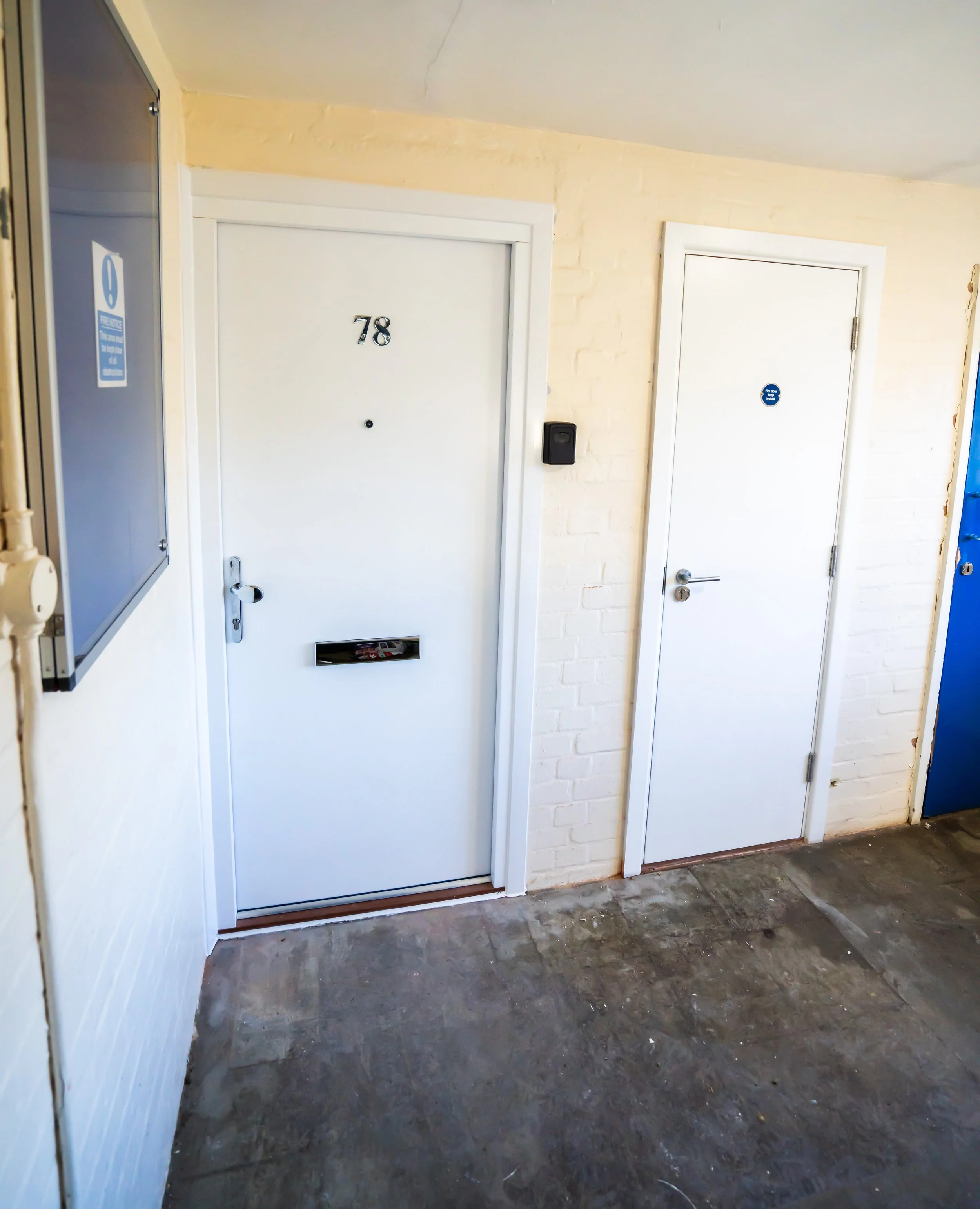 A hallway with two white doors, one numbered 78 and the other with a sign indicating fire door, a black keypad or lock on the wall, and an electrical box on the left. The floor is dark and slightly worn, with some paint or debris scattered.