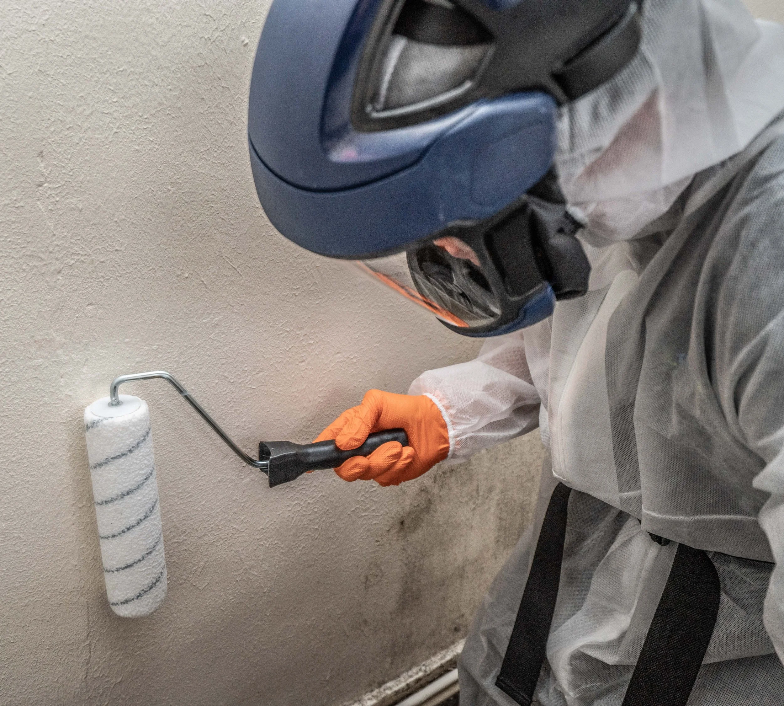 Person wearing a white protective suit, orange gloves, a black face mask, and a blue helmet, painting a wall with a paint roller.