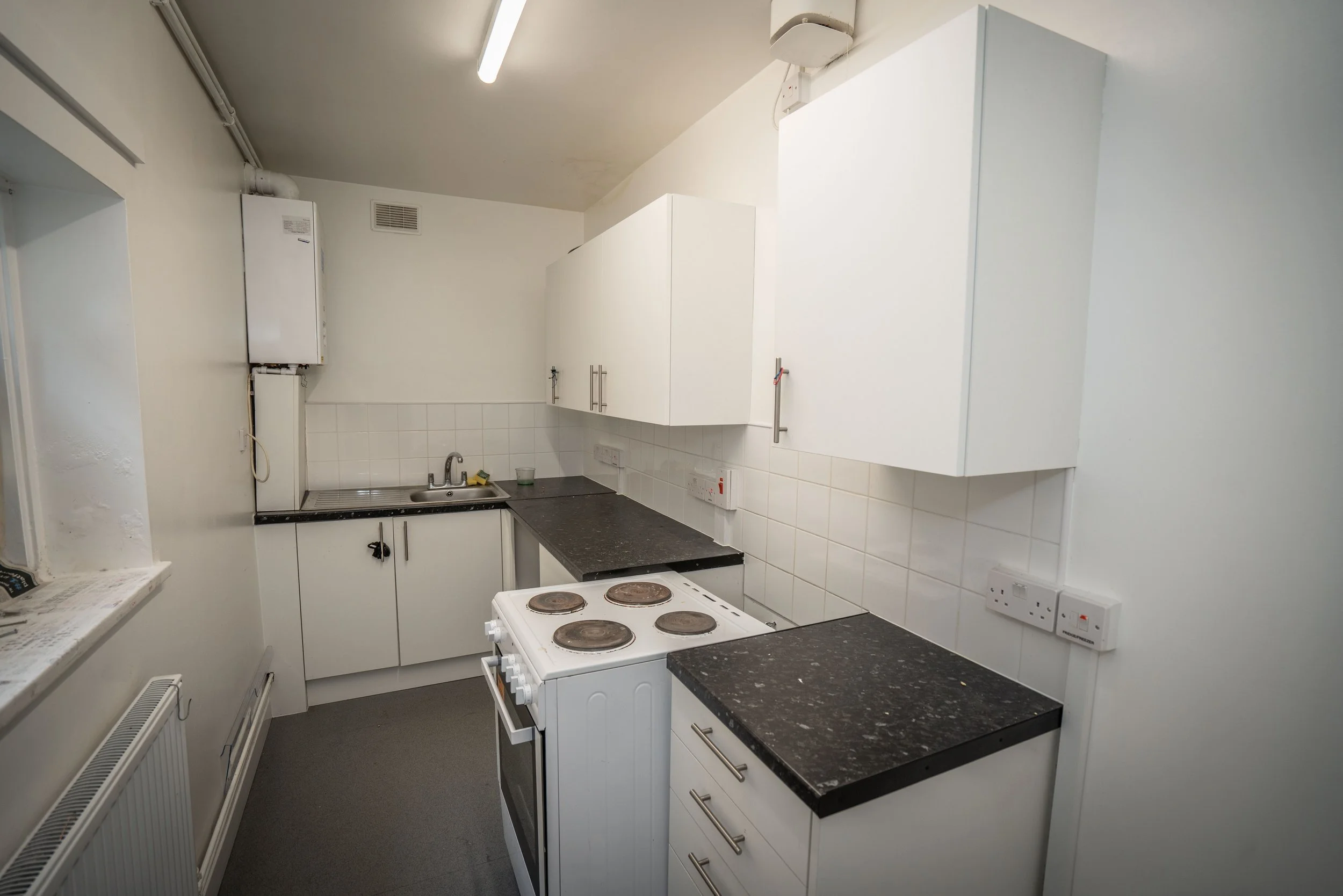 Small kitchen with white cabinets, black speckled countertops, a white stove with four burners, a sink with a metal faucet, and a wall-mounted white boiler. There is a radiator along the bottom of the left wall, and electrical outlets on the backspla