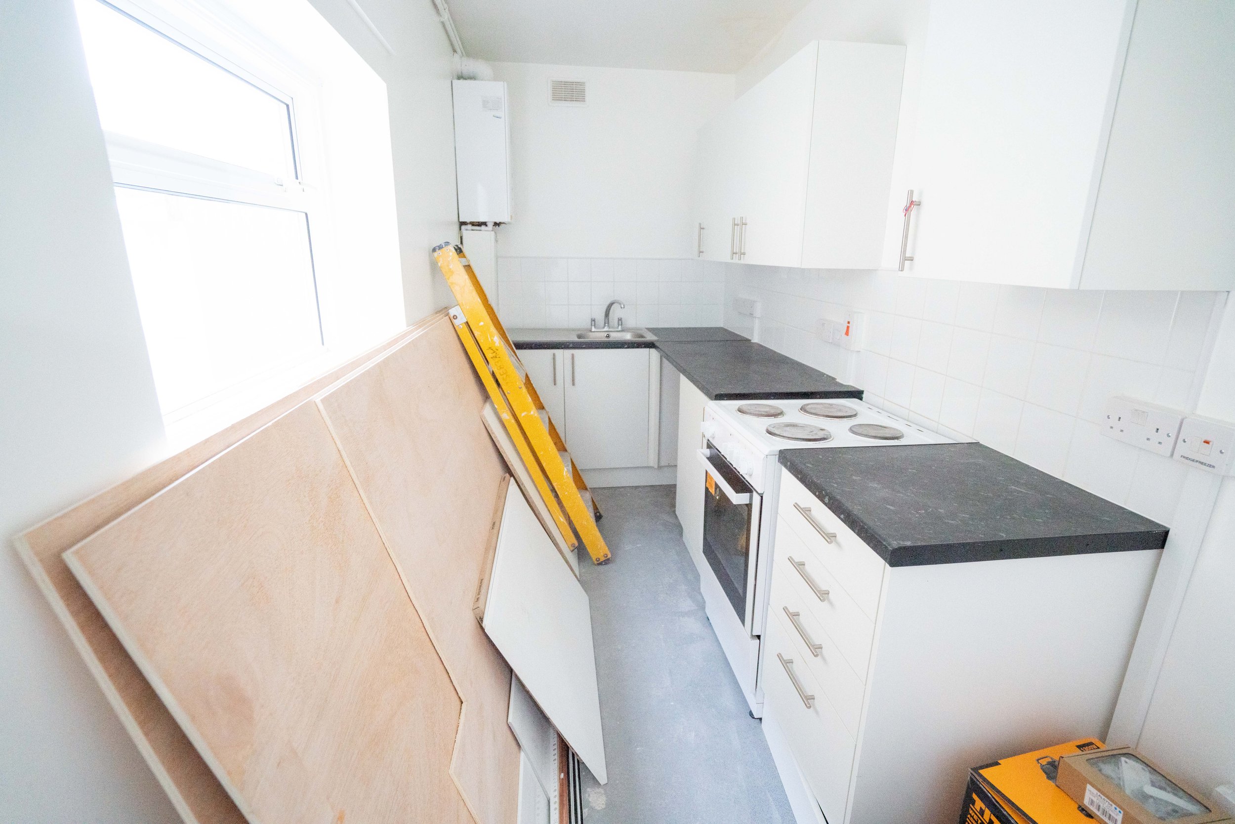 Empty kitchen with white cabinets, black countertop, stove, sink, a yellow ladder, and a large piece of wood leaning against the wall by the window.