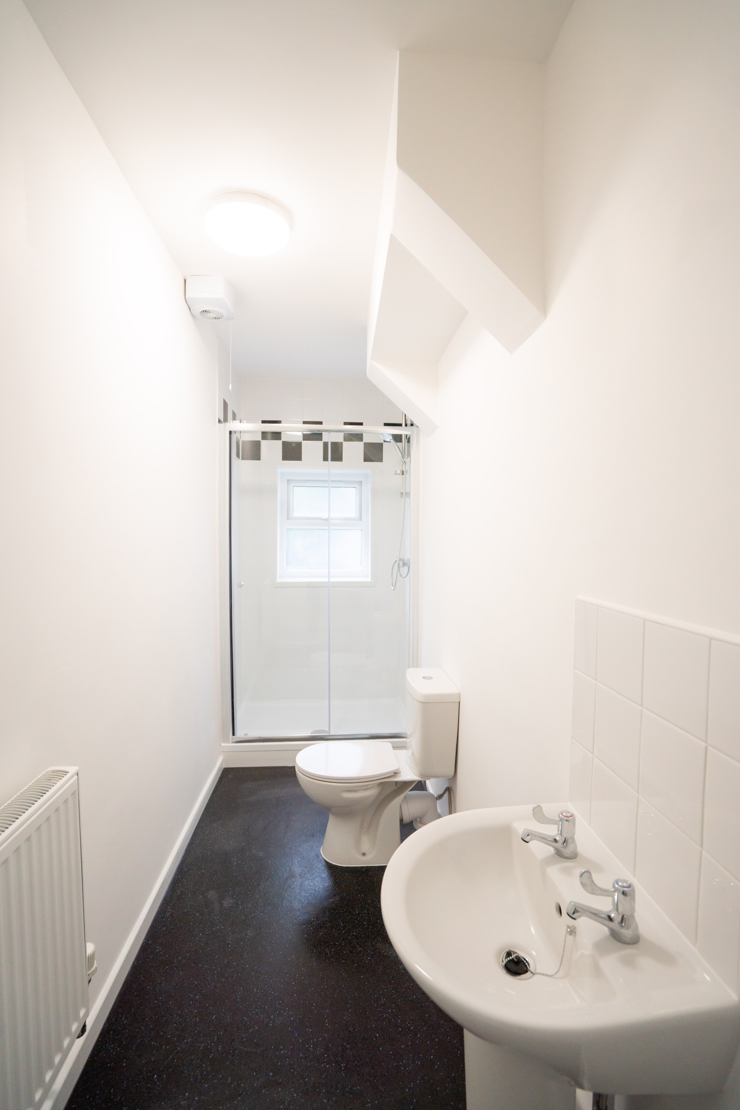 A small bathroom with white walls, featuring a toilet next to a sink with dual faucets, a window above the shower, and a shower with sliding glass doors. There is a radiator on the left wall and a black speckled floor.
