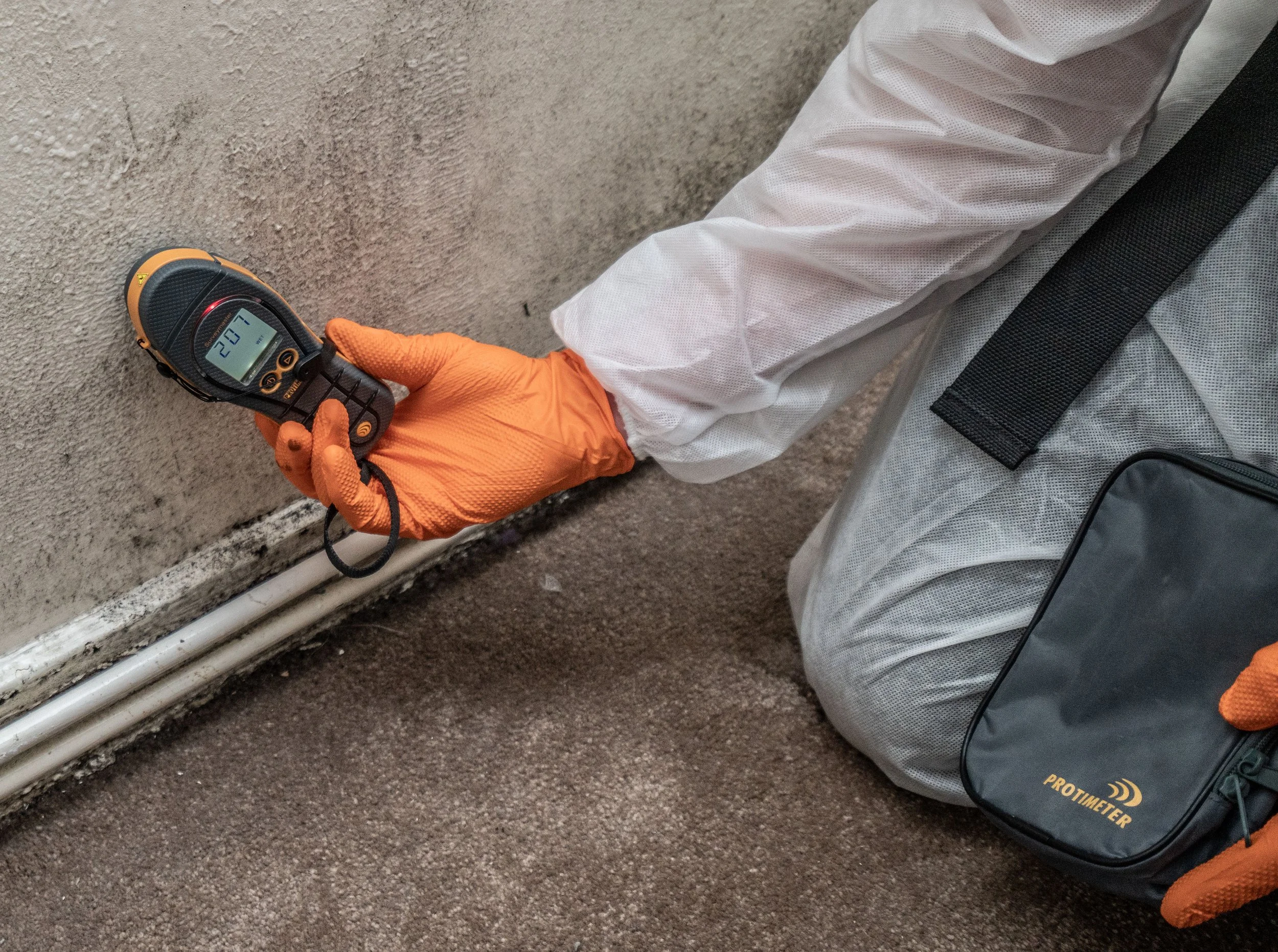 A person wearing an orange glove holding a handheld device with a digital display showing the number 20, while sitting on a gray textured carpet near white pipes and a gray and black backpack.