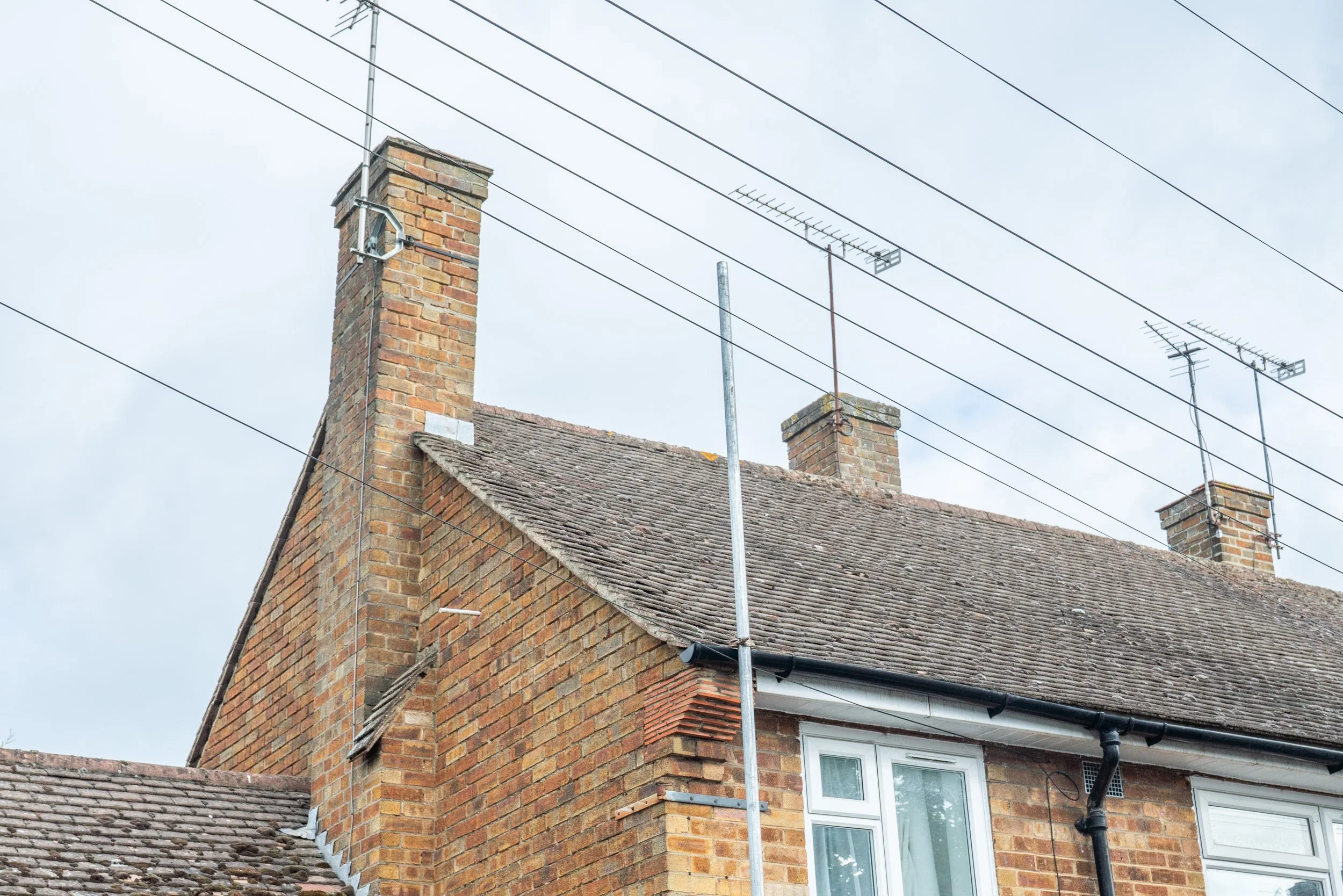 Close-up of a brick house roof with chimneys and TV antennas against a cloudy sky.
