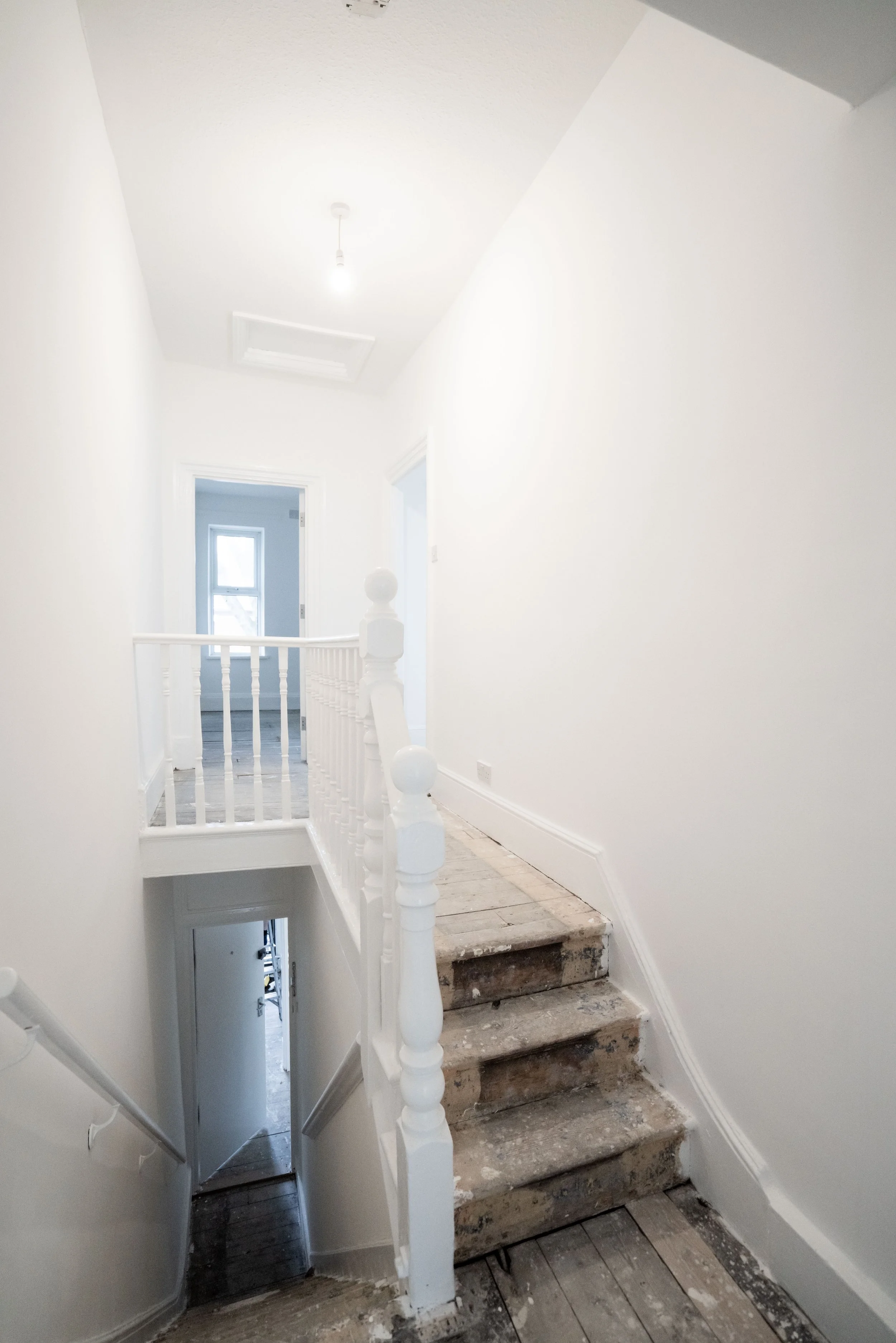 Interior of a house under renovation showing a staircase with marble steps, white railing, and a hallway with open doors, white walls, a ceiling with a light bulb, and a window in the background.