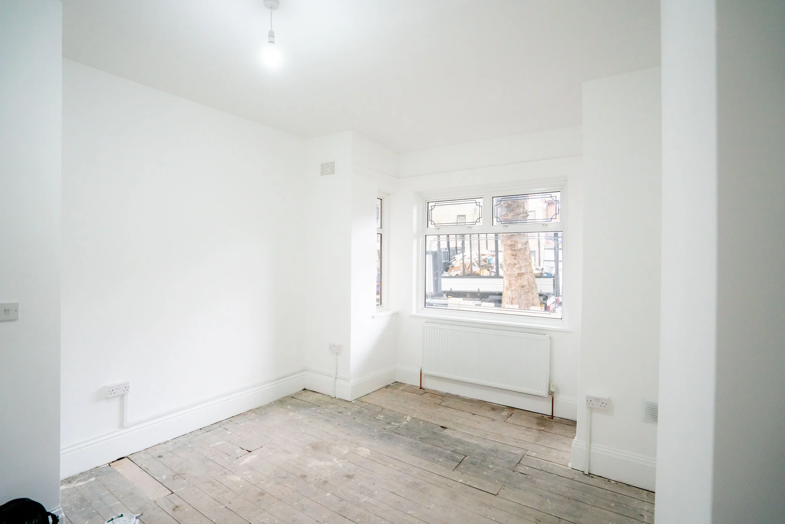 An empty room with white walls and large windows, indicating it is under renovation or construction, with visible floorboards and some construction materials on the floor.