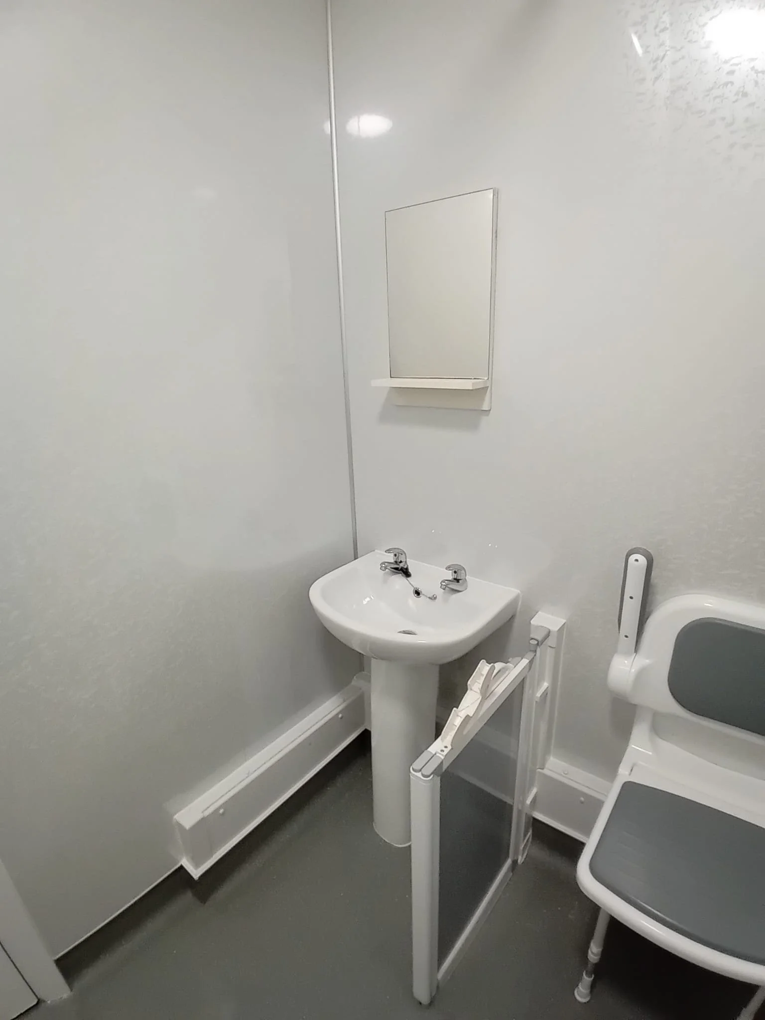 A hospital bathroom with a white sink and a gray and white toilet, a mirror above the sink, and a white privacy screen divider.