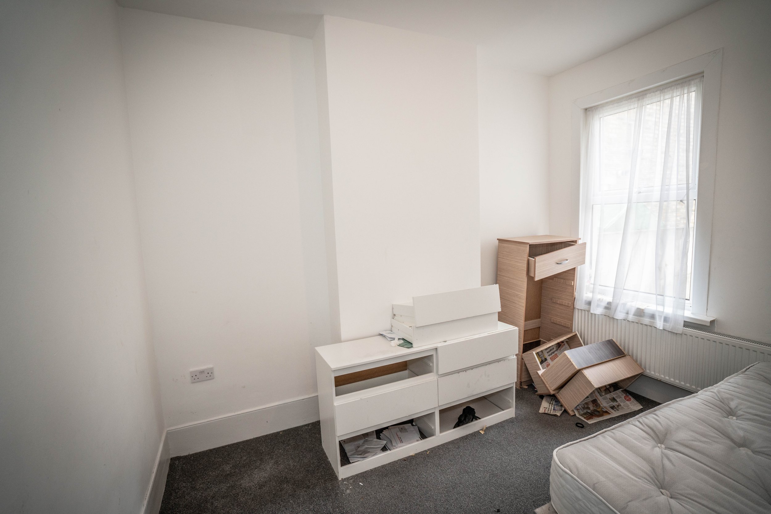 A room with white walls, a large window with sheer curtains, a white storage unit, a cardboard box, and some papers on the floor.