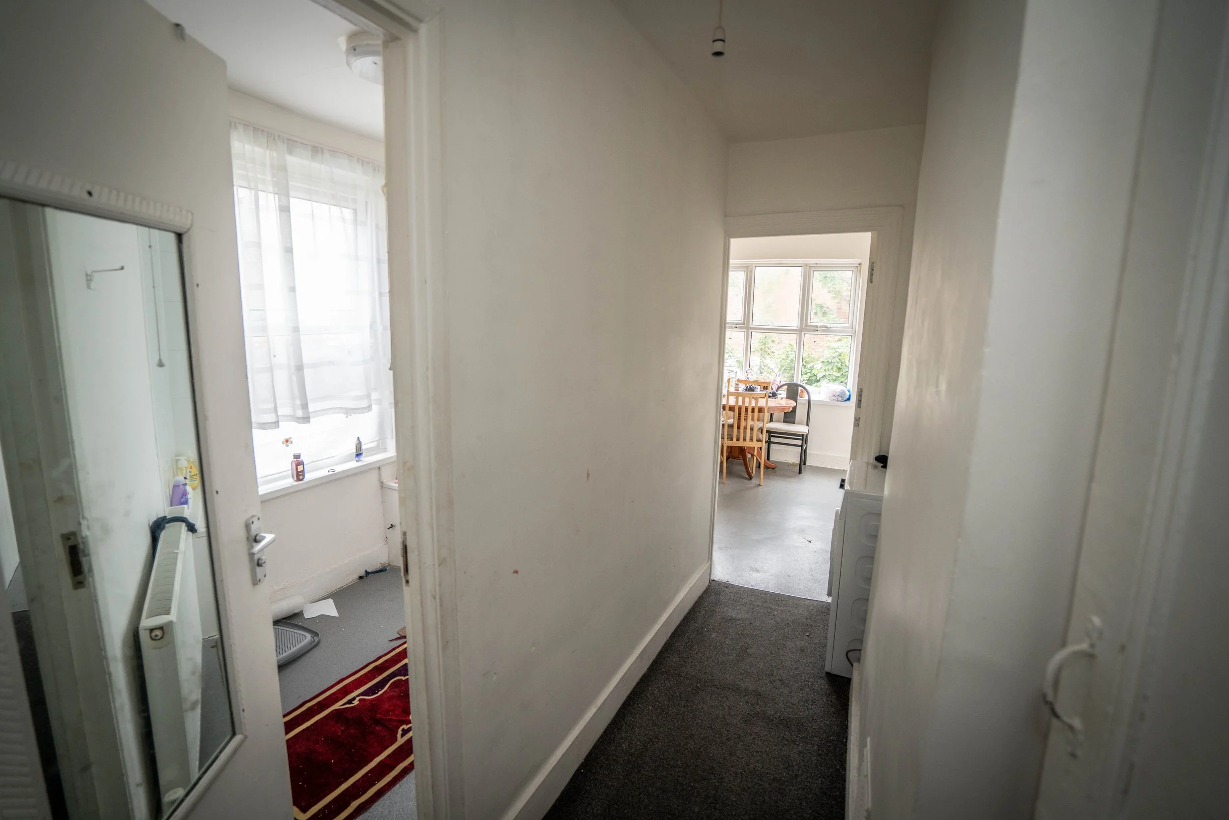 View of an apartment interior with a hallway leading to a dining area with a window. The hallway has dark carpeting, white walls, and a small section of a white washing machine is visible.