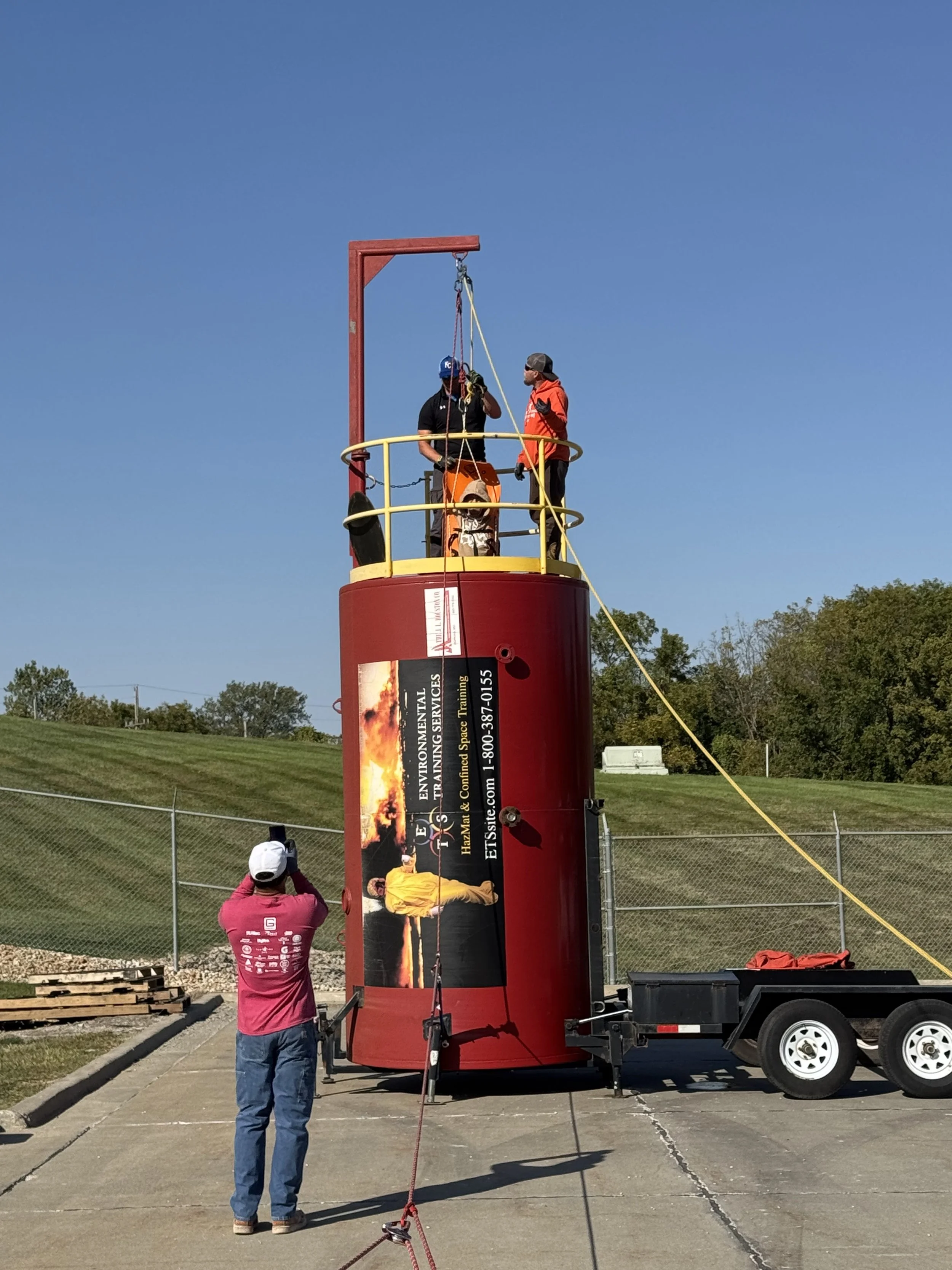 Two workers standing inside a yellow safety railing on top of a large red environmental training tank, preparing for a safety drill. One worker is adjusting equipment while another observes. A person in a pink shirt takes a photo. The scene is outdoors with a blue sky and grassy area beyond a chain-link fence.