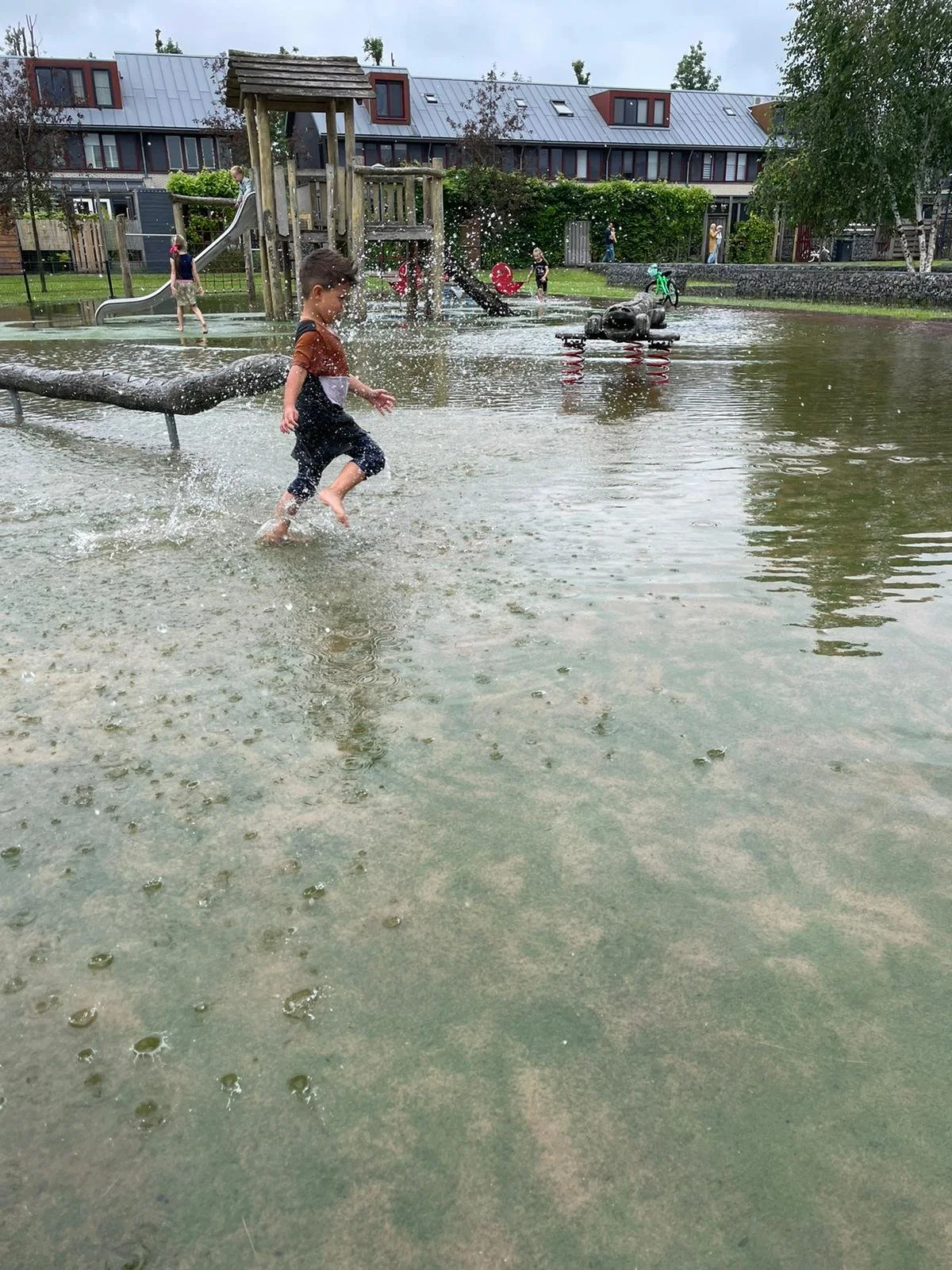 Children playing in a flooded playground, splashing water and walking through shallow water, with playground equipment and houses in the background.