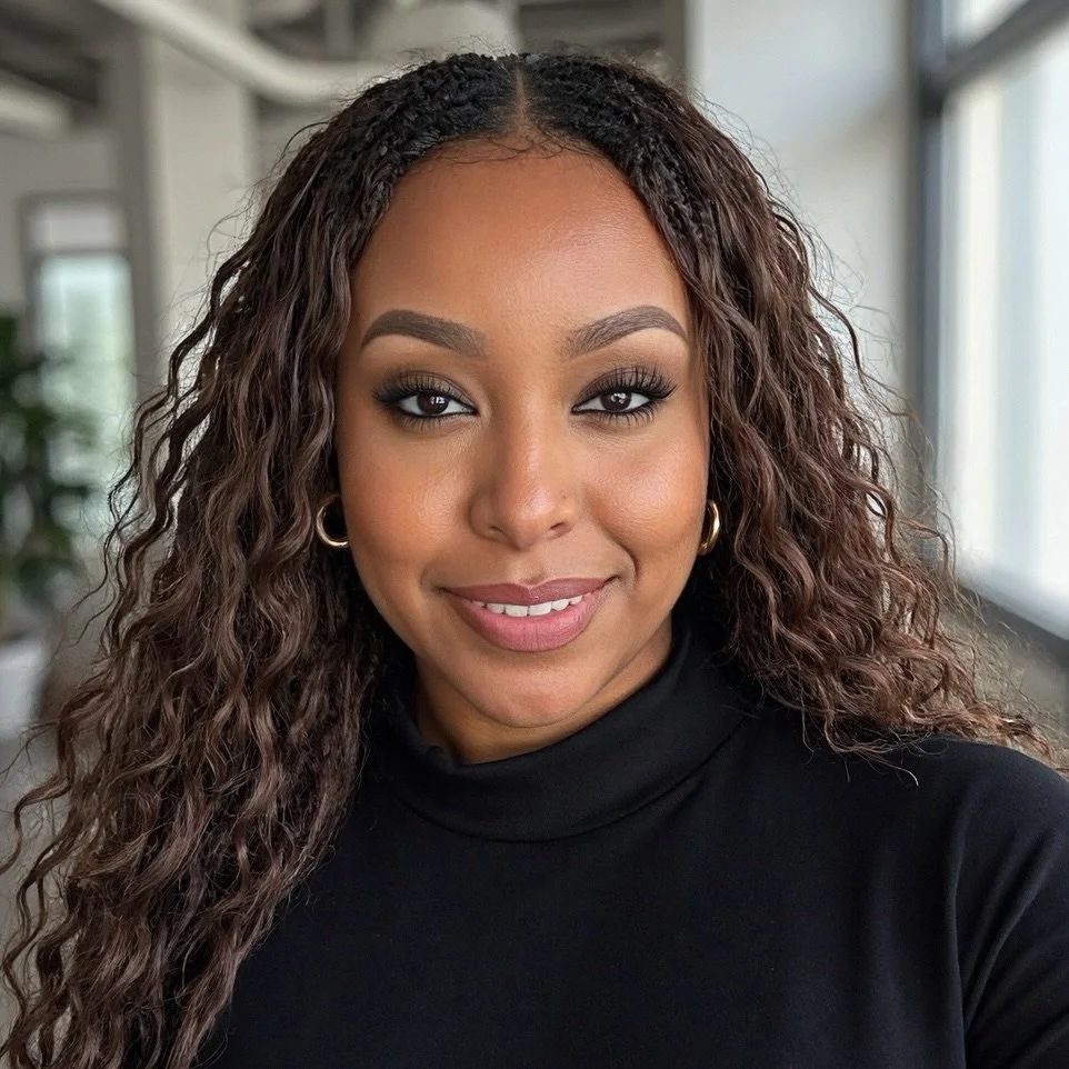 Close-up of a woman with long curly hair, wearing a black turtleneck and gold hoop earrings, smiling in a well-lit indoor setting.
