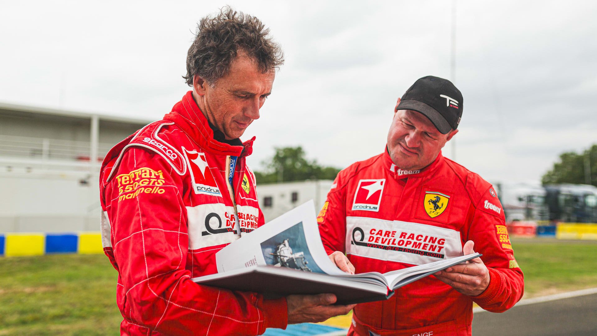 Two race car drivers in red racing suits reviewing a book at a race track.
