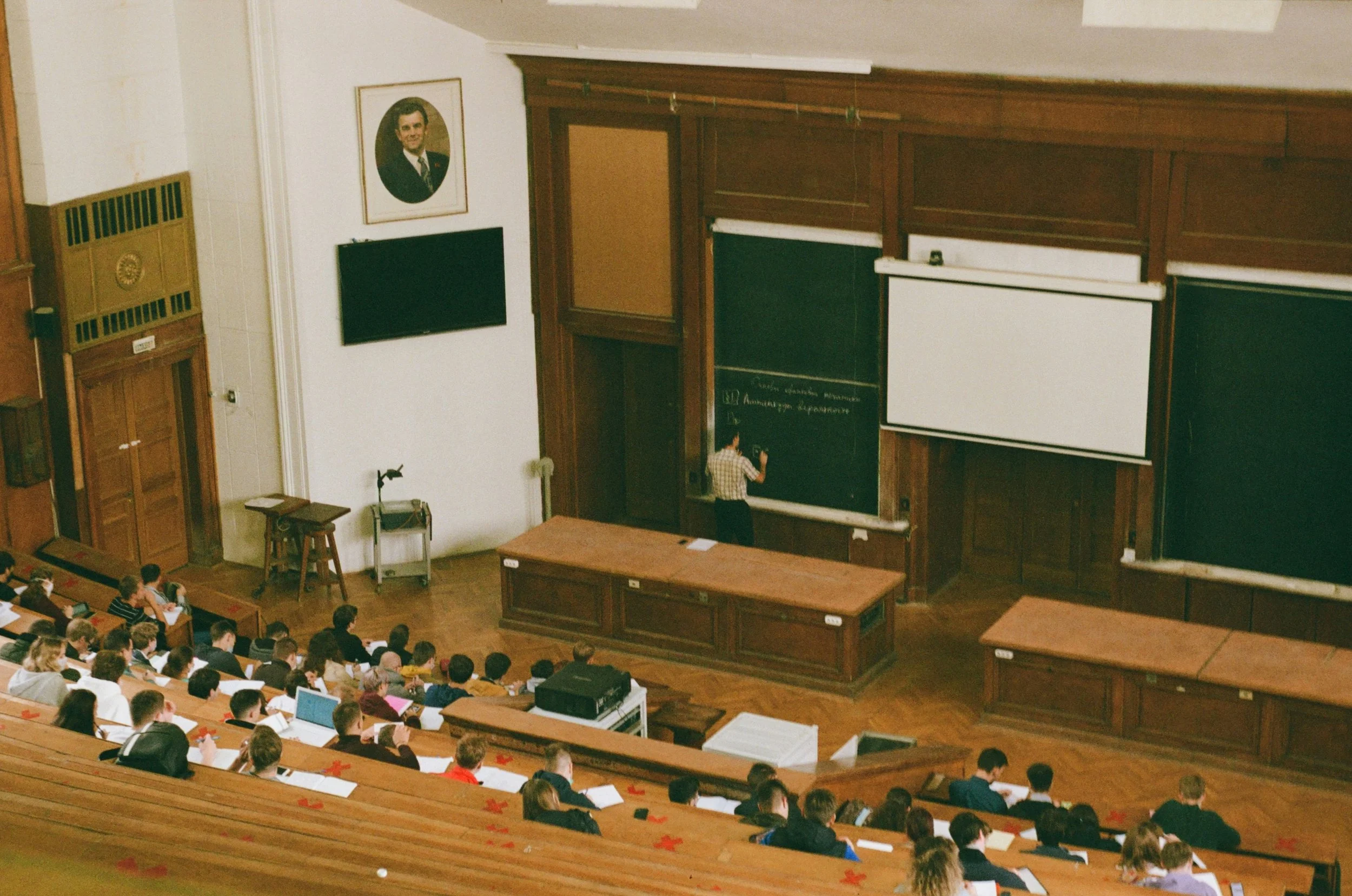 A university lecture hall filled with students, with a teacher writing on a blackboard at the front. The classroom features dark wood paneling and a portrait hanging on the wall.
