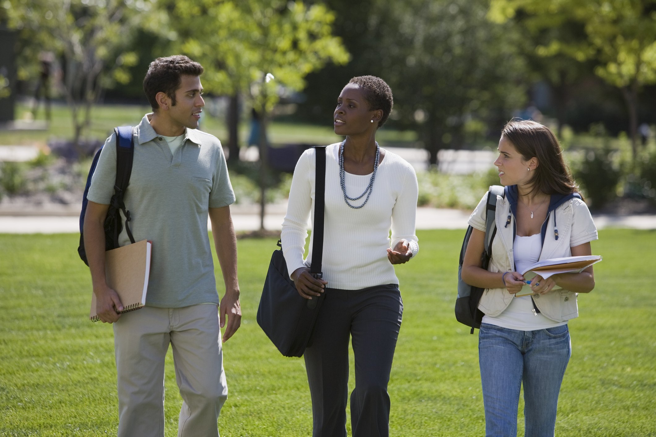 Two students carrying books and backpacks walking outdoors near modern buildings and trees on a sunny day.