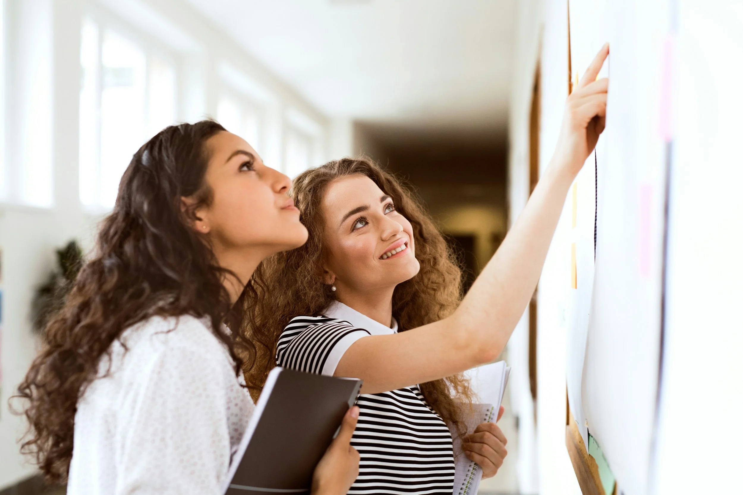 Two young women with curly hair looking at a wall with sticky notes or papers, one is writing or pointing, holding notebooks and papers, in a bright hallway.