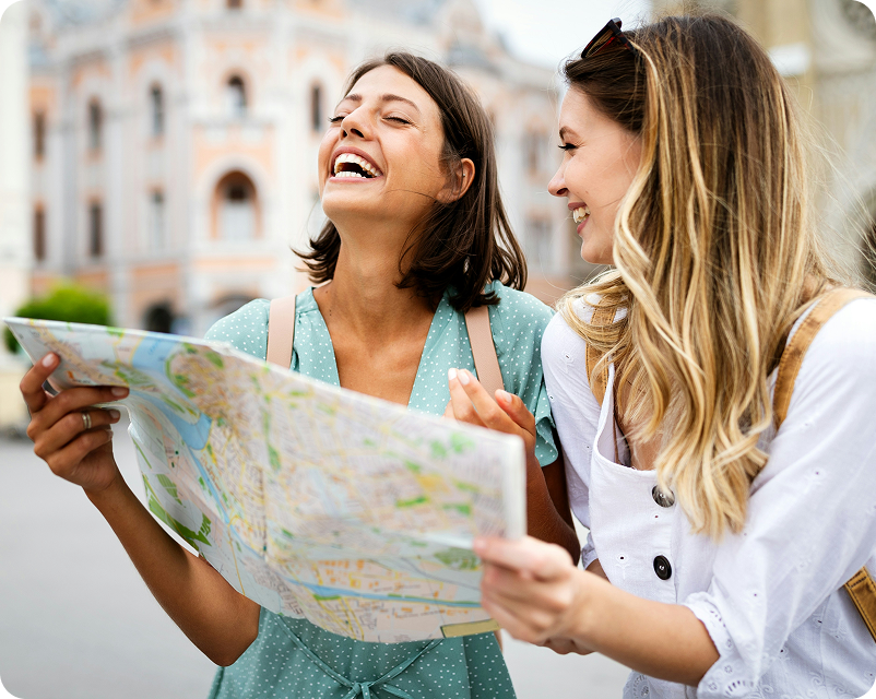 Two young women are looking at a map and smiling while sightseeing outdoors in front of a historic building.
