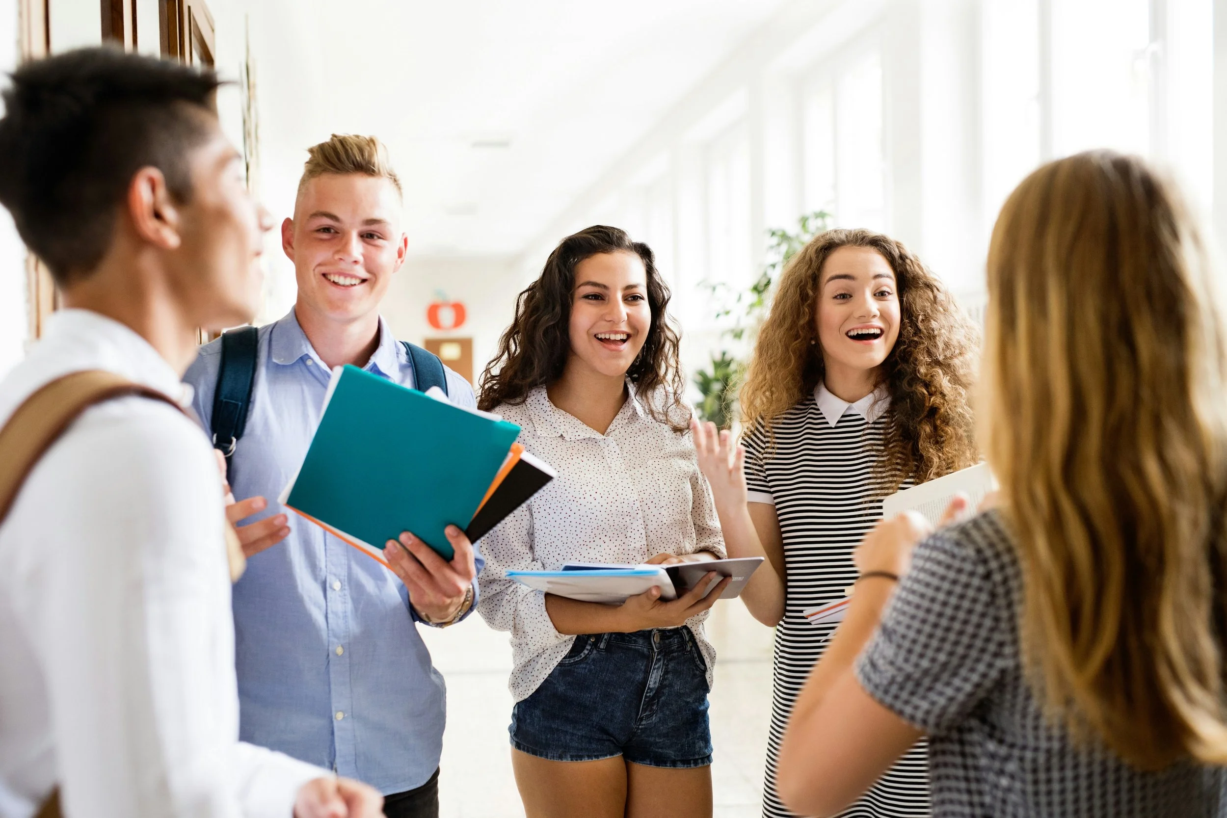 Group of five diverse teenagers talking and smiling in school hallway, holding notebooks and papers.