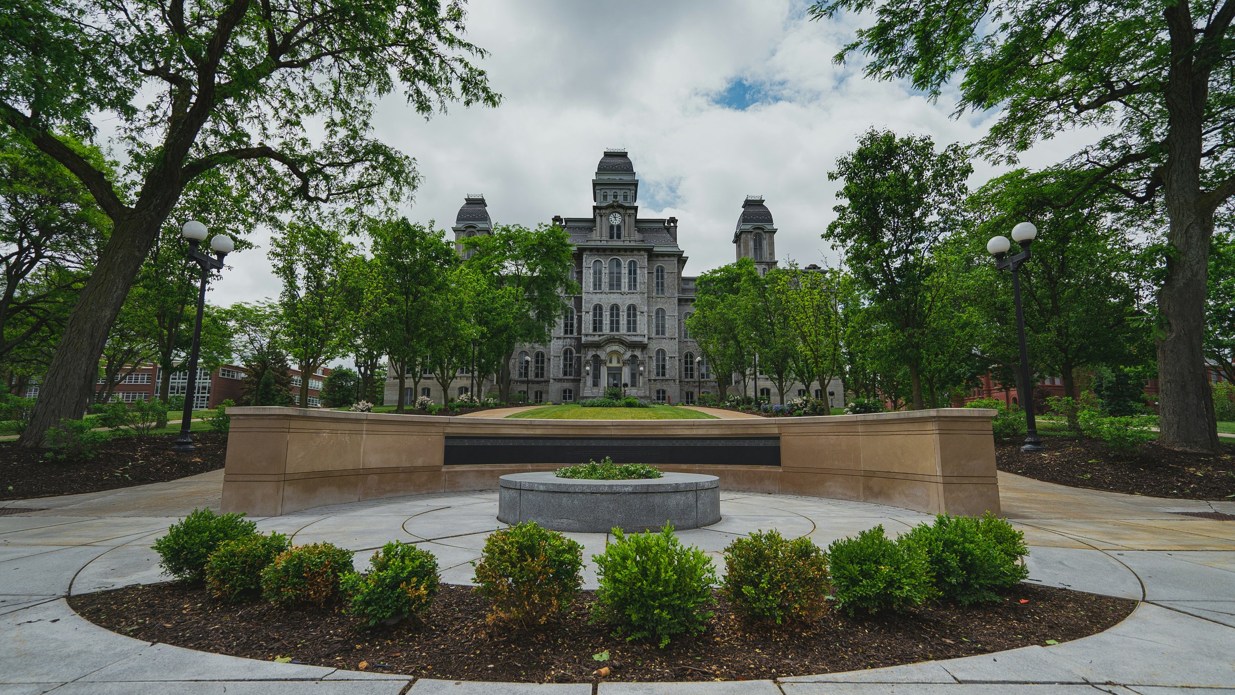 A historic gray stone building with a clock tower, surrounded by green trees, with a paved circular walkway and bushes in the foreground.