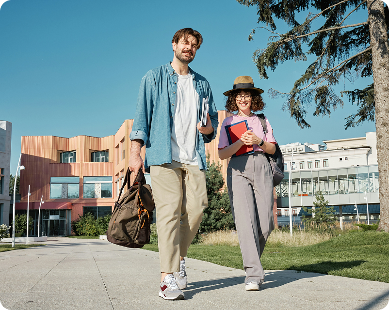 Two students carrying books and backpacks walking outdoors near modern buildings and trees on a sunny day.