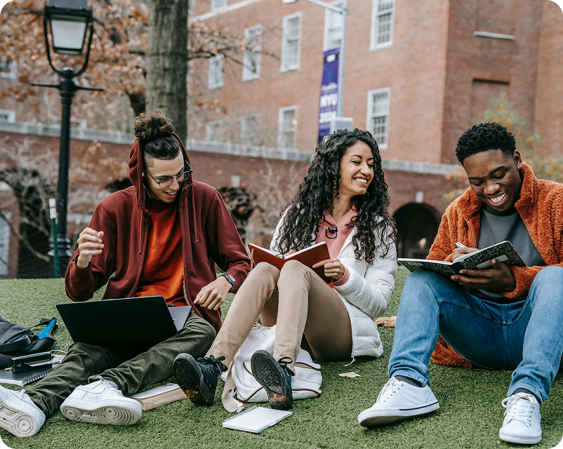 Three college students sitting on grass outside, studying and laughing, with notebooks and laptops.