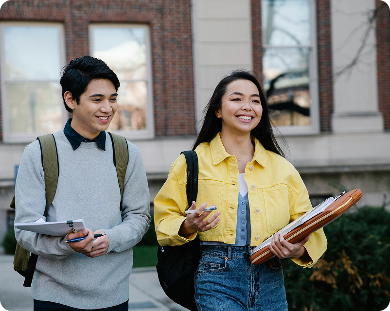 Three college students sitting on grass outdoors, laughing and studying with laptops and notebooks.