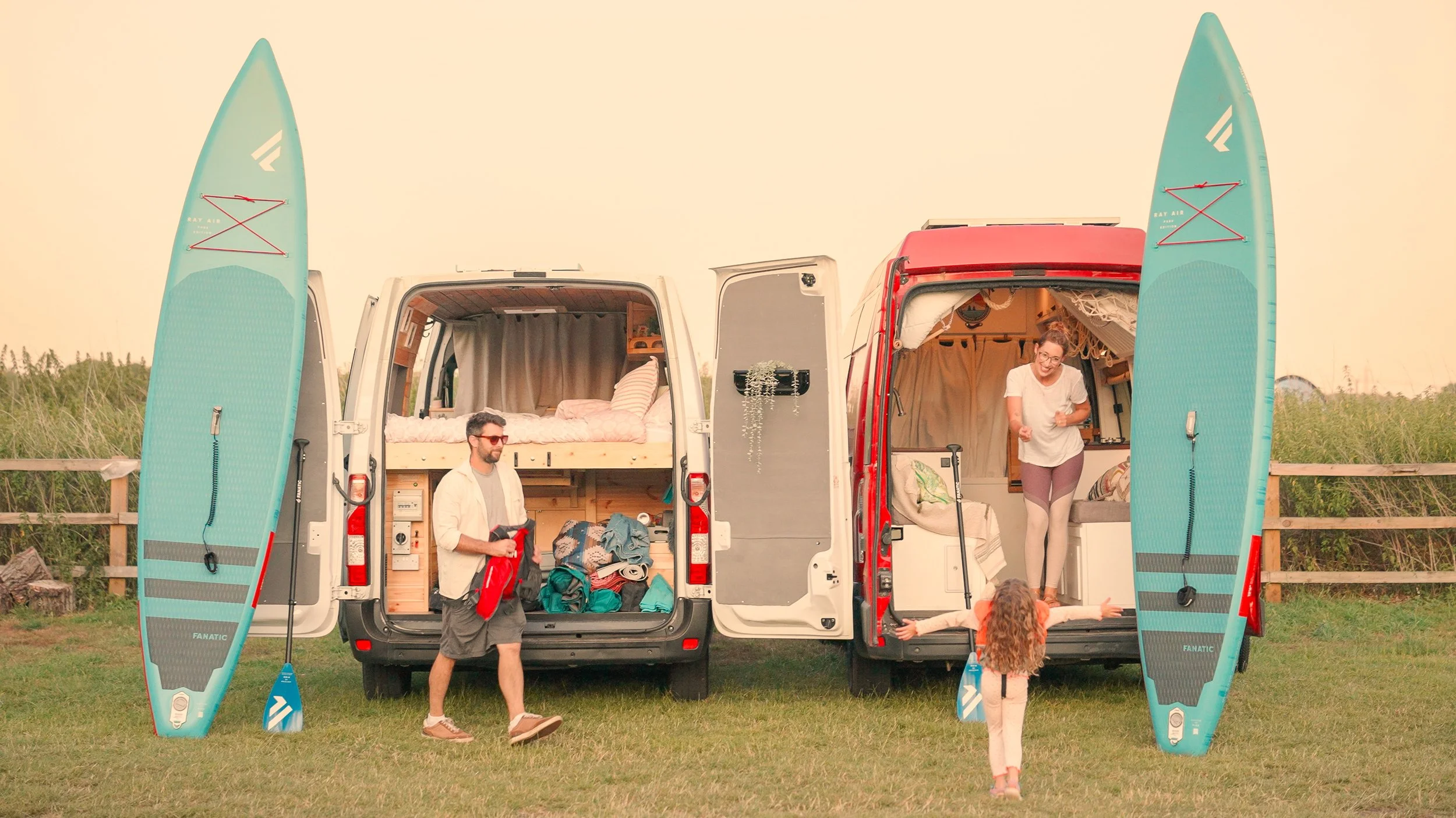 Family setting up a camping van with paddleboards on a grassy field at sunset, with three people, a man carrying a paddle, and a woman and child near the van.