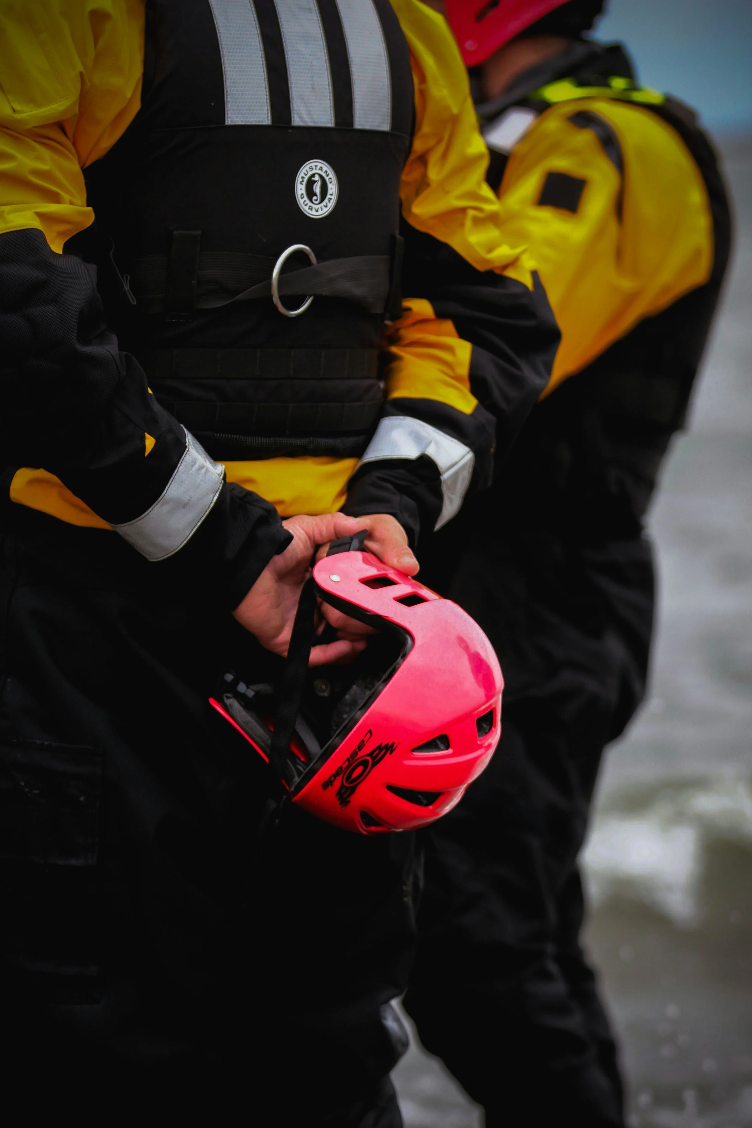Person wearing a yellow and black waterproof jacket holding a pink safety helmet, with additional people in similar attire in the background by water.