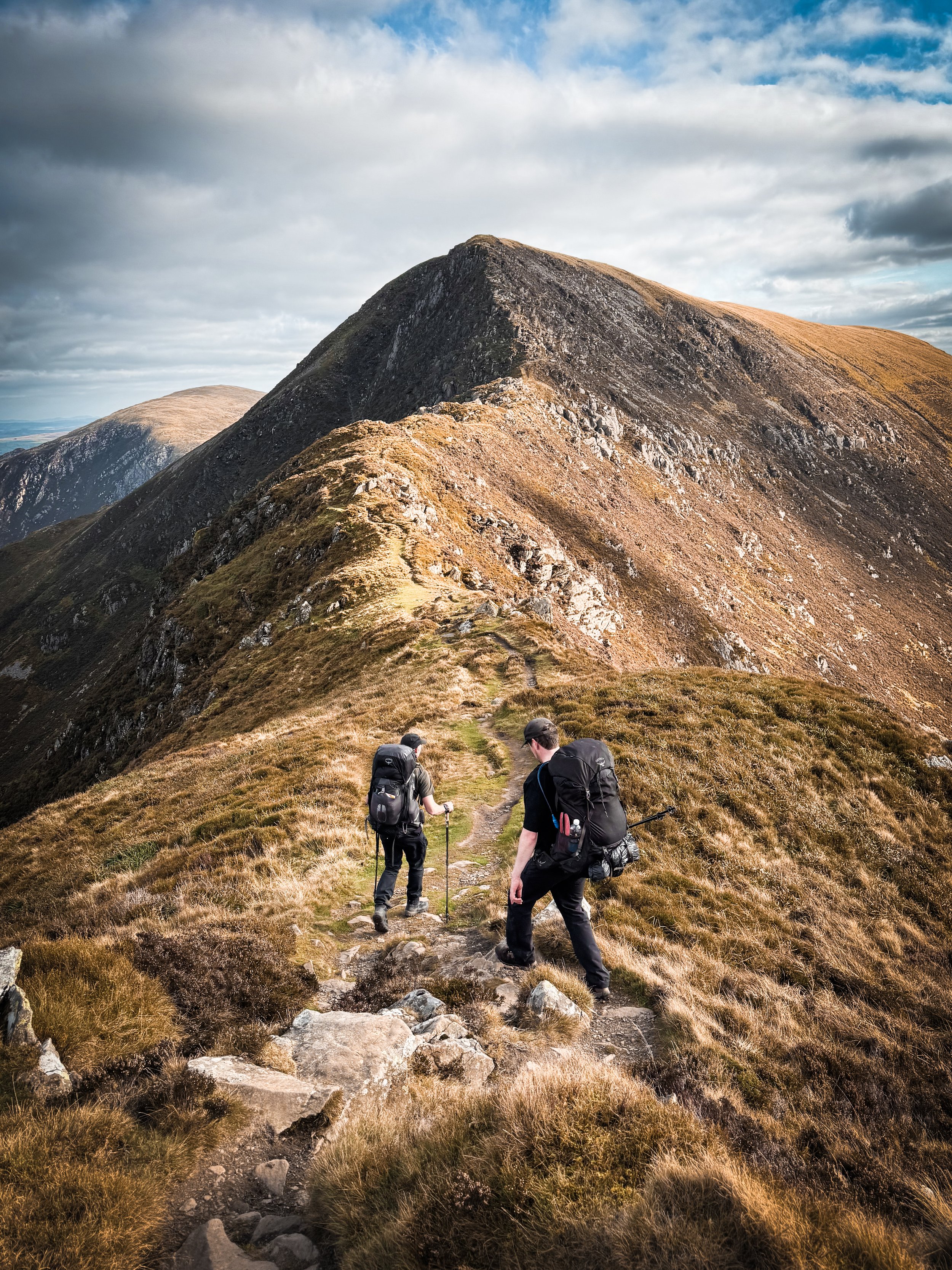 Two hikers carrying backpacks and trekking poles ascending a mountain trail with grassy and rocky terrain, leading toward a mountain peak under a partly cloudy sky.