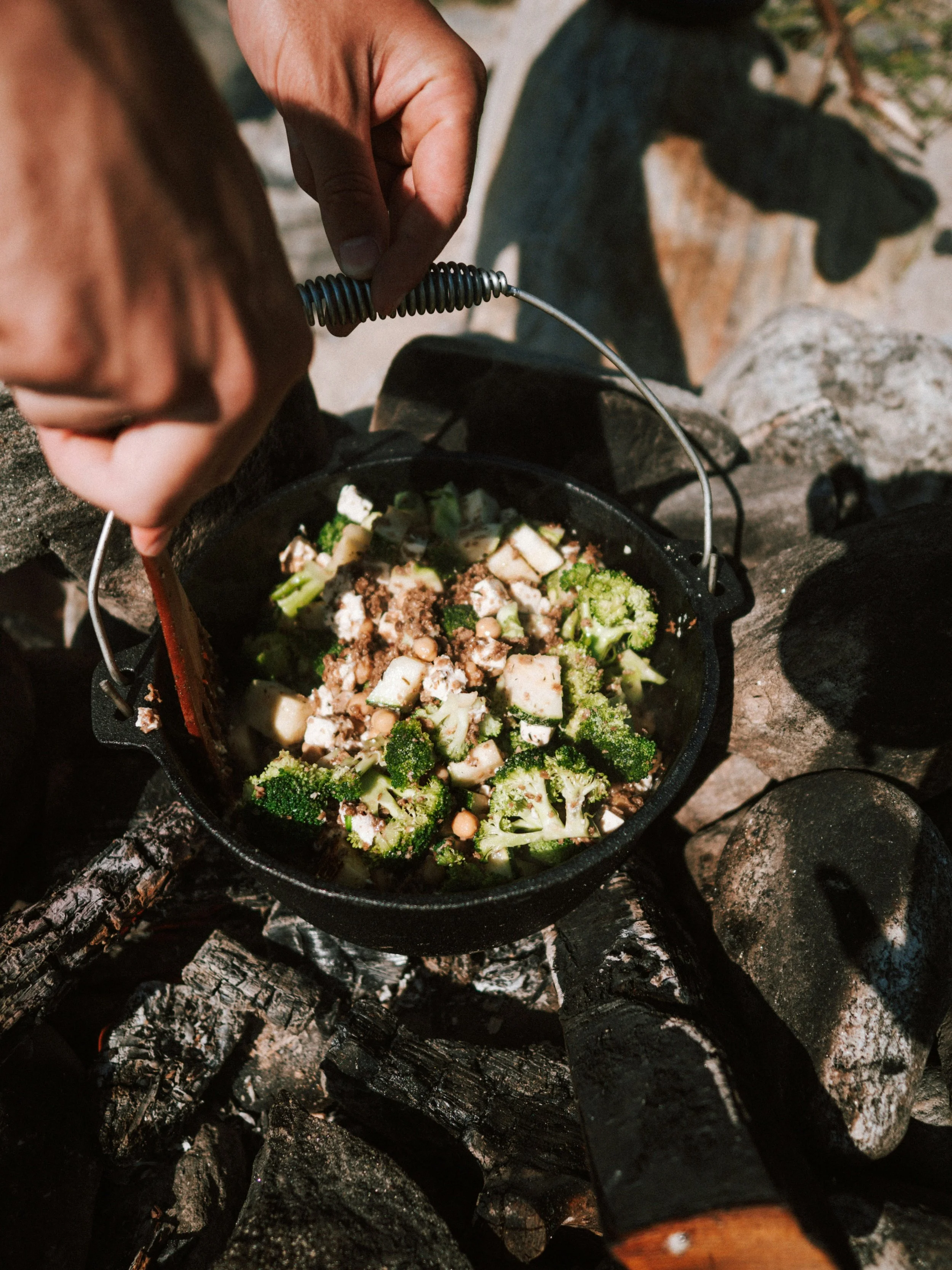 Person cooking vegetables in a cast iron pot over an outdoor campfire.