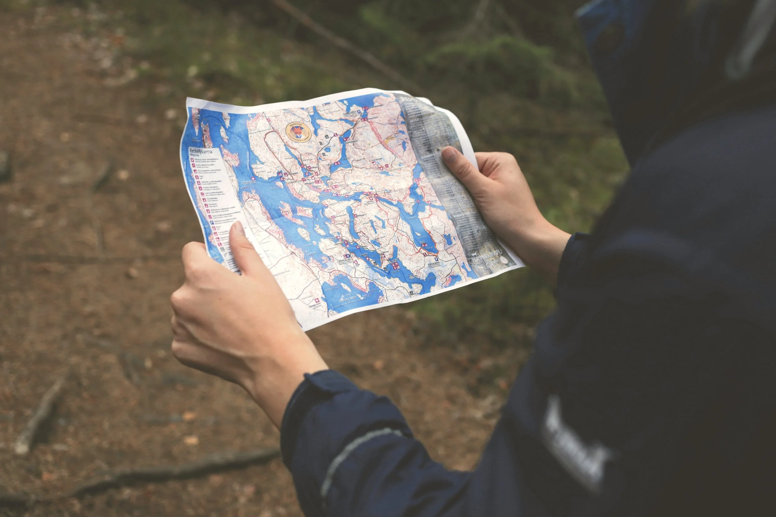 Person in outdoor clothing holding and reading a detailed map of lakes and waterways, standing on a dirt forest trail.