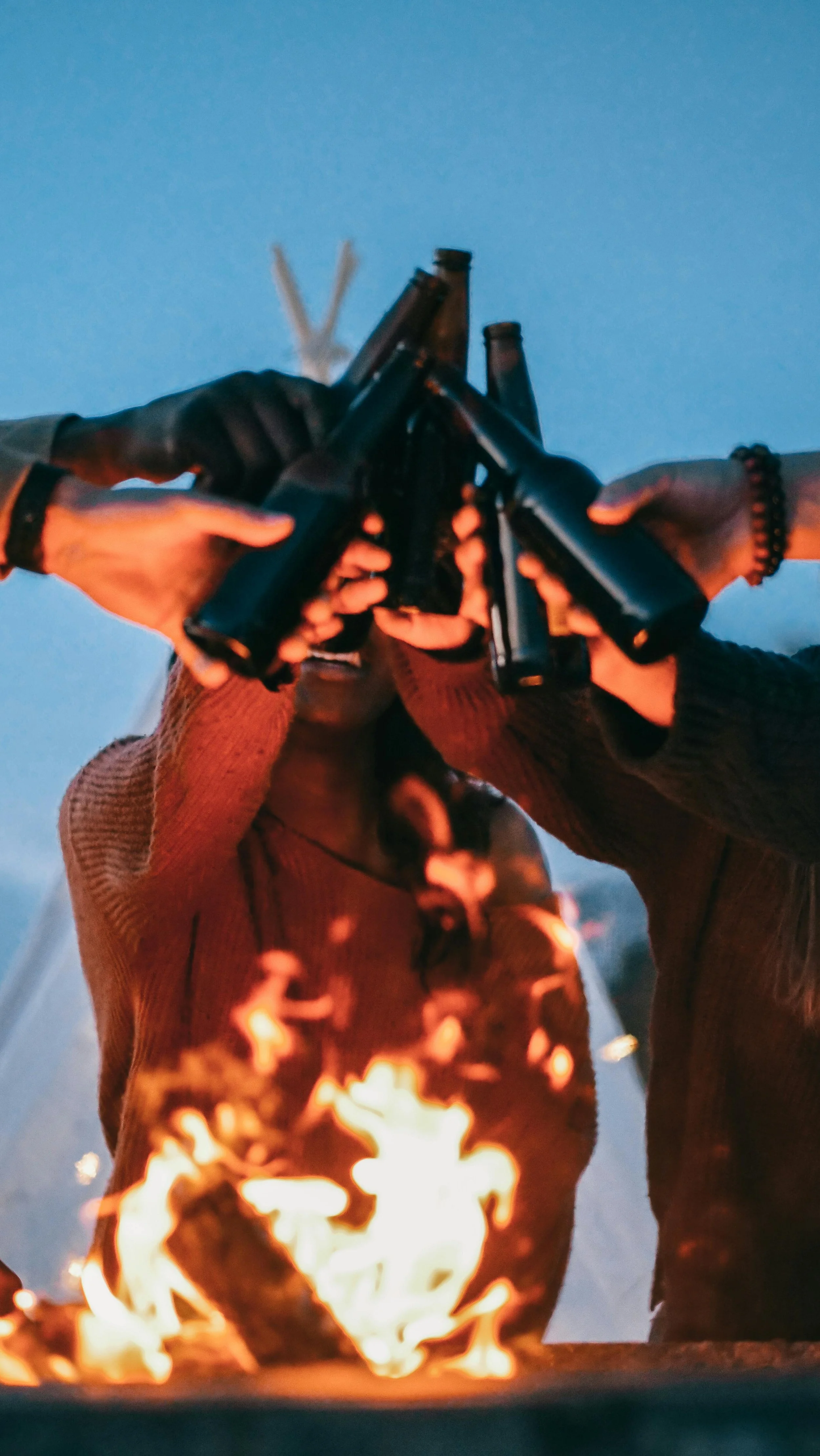 Group of people toasting with beer bottles over a fire during the evening.