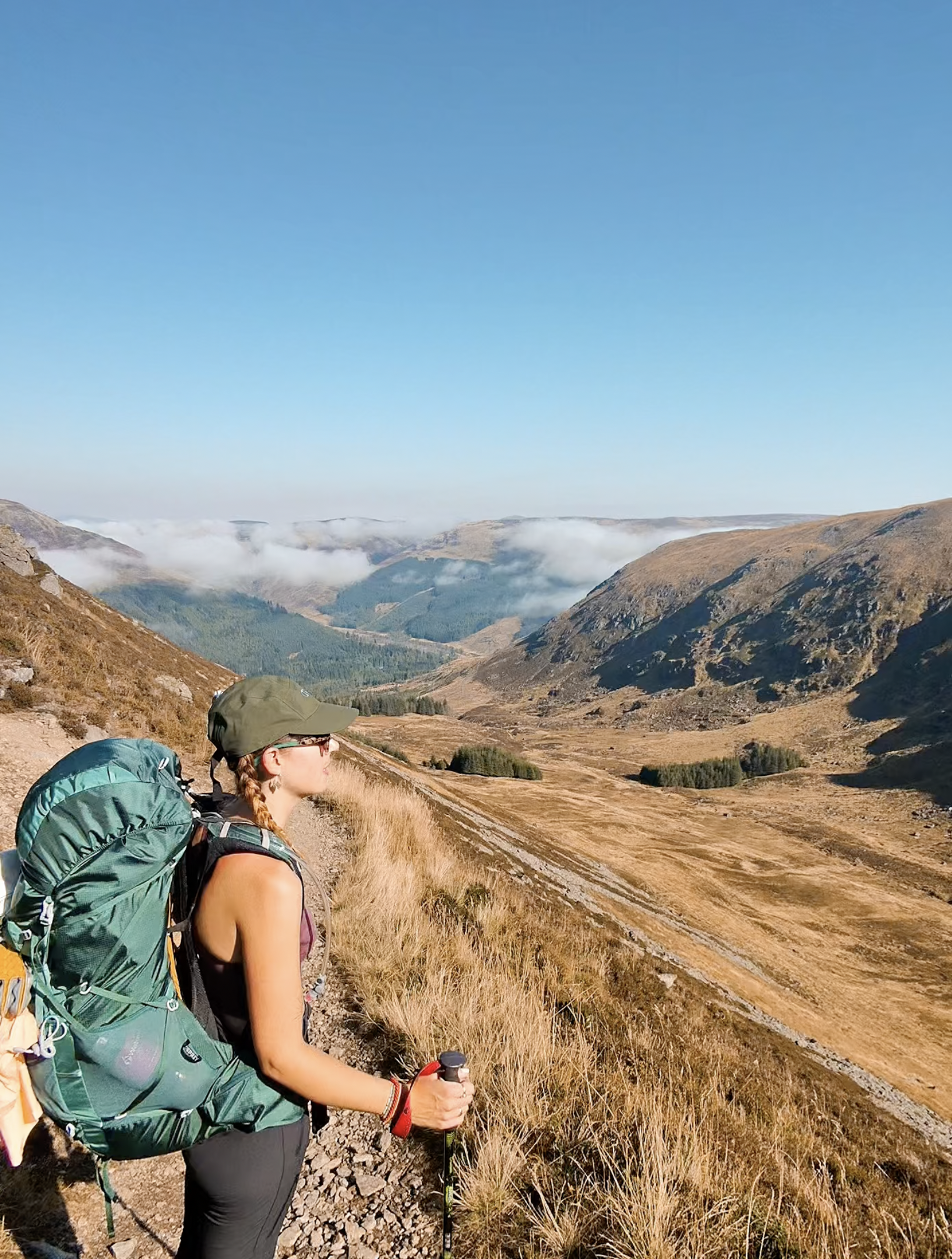 A woman hiking on a trail in a mountainous landscape with dry grass, rolling hills, and a blue sky.
