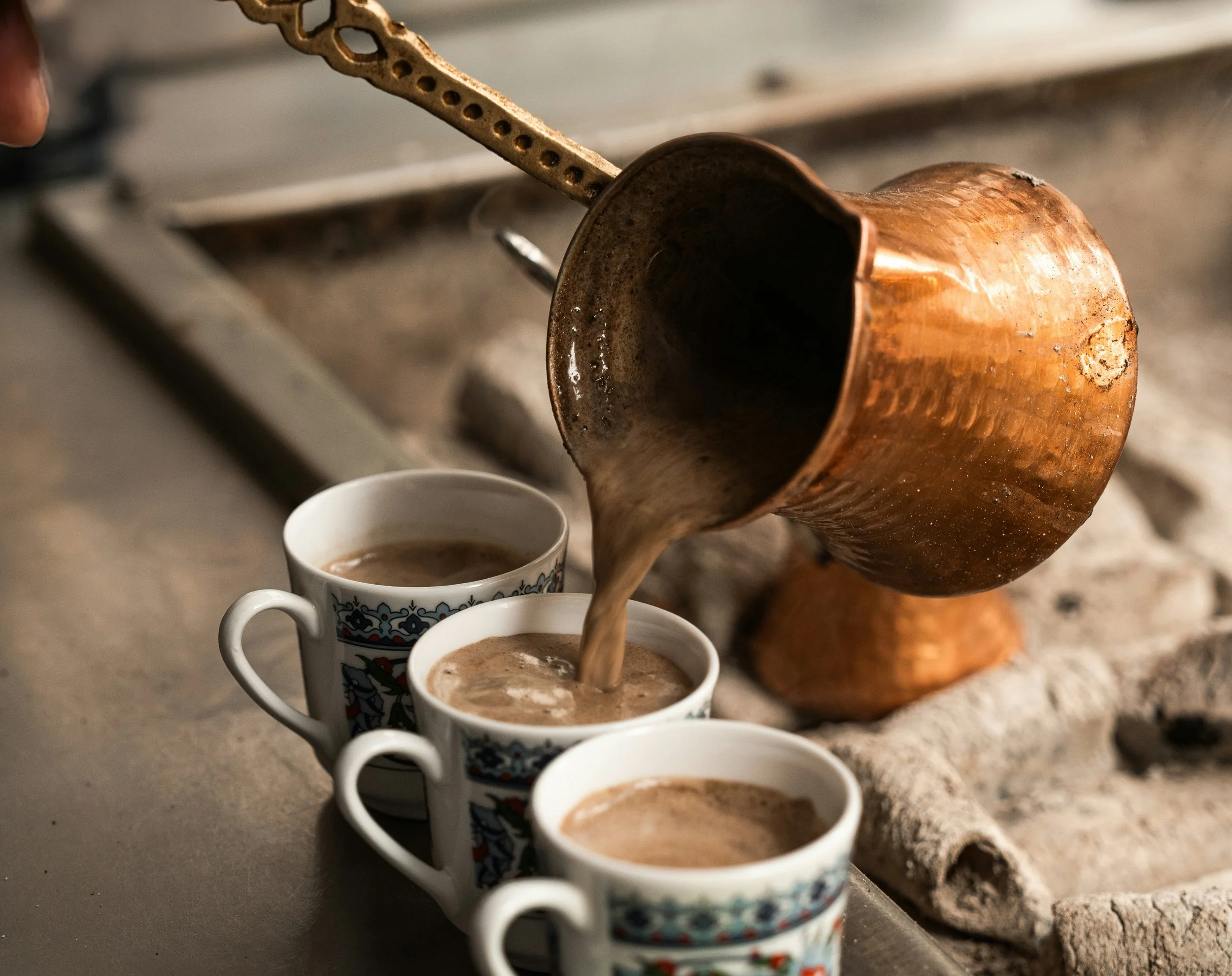 Pouring coffee from a traditional copper cezve into three patterned ceramic cups