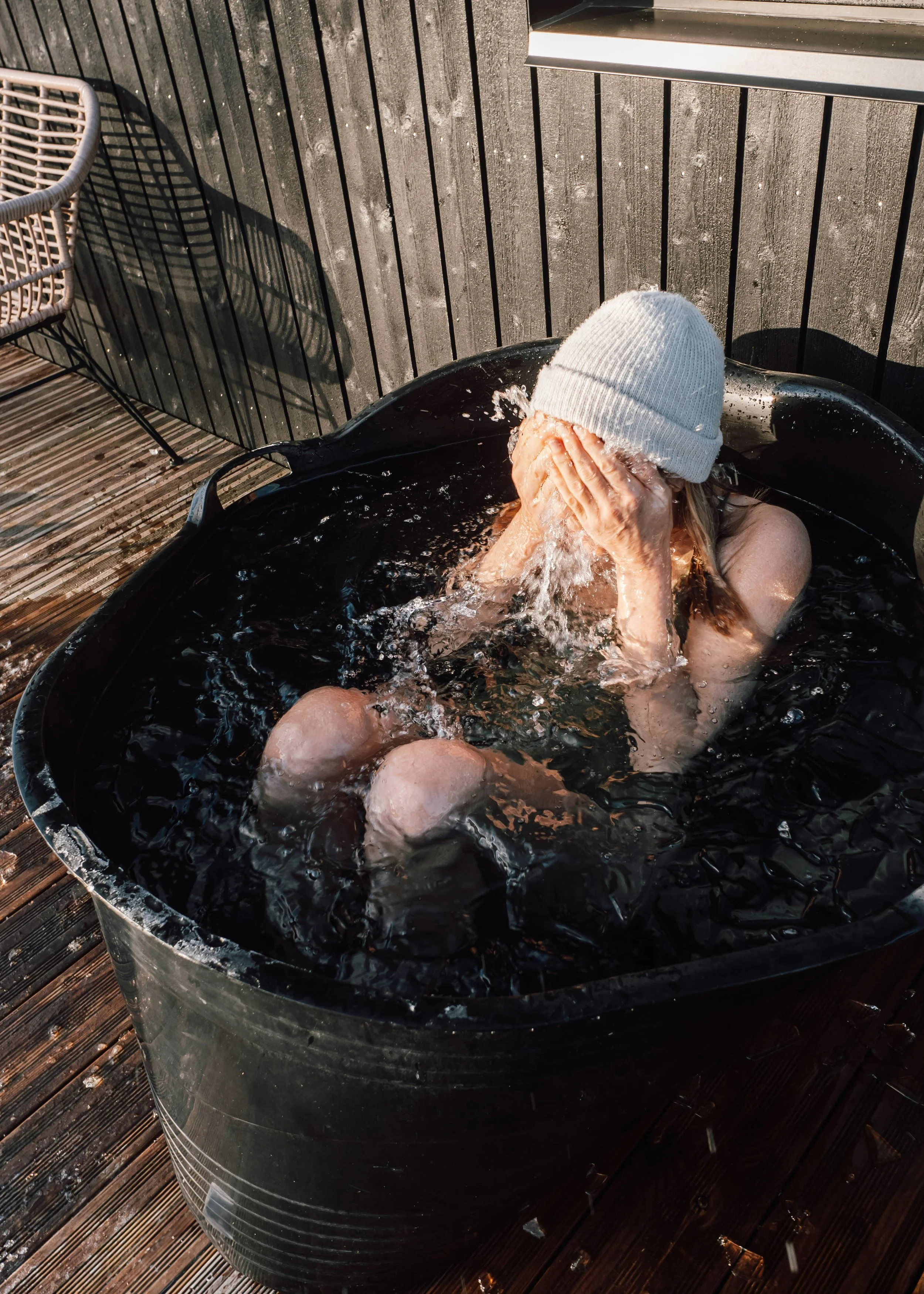 A woman wearing a white beanie is sitting in a black bucket of water on a wooden deck, splashing water on her face.