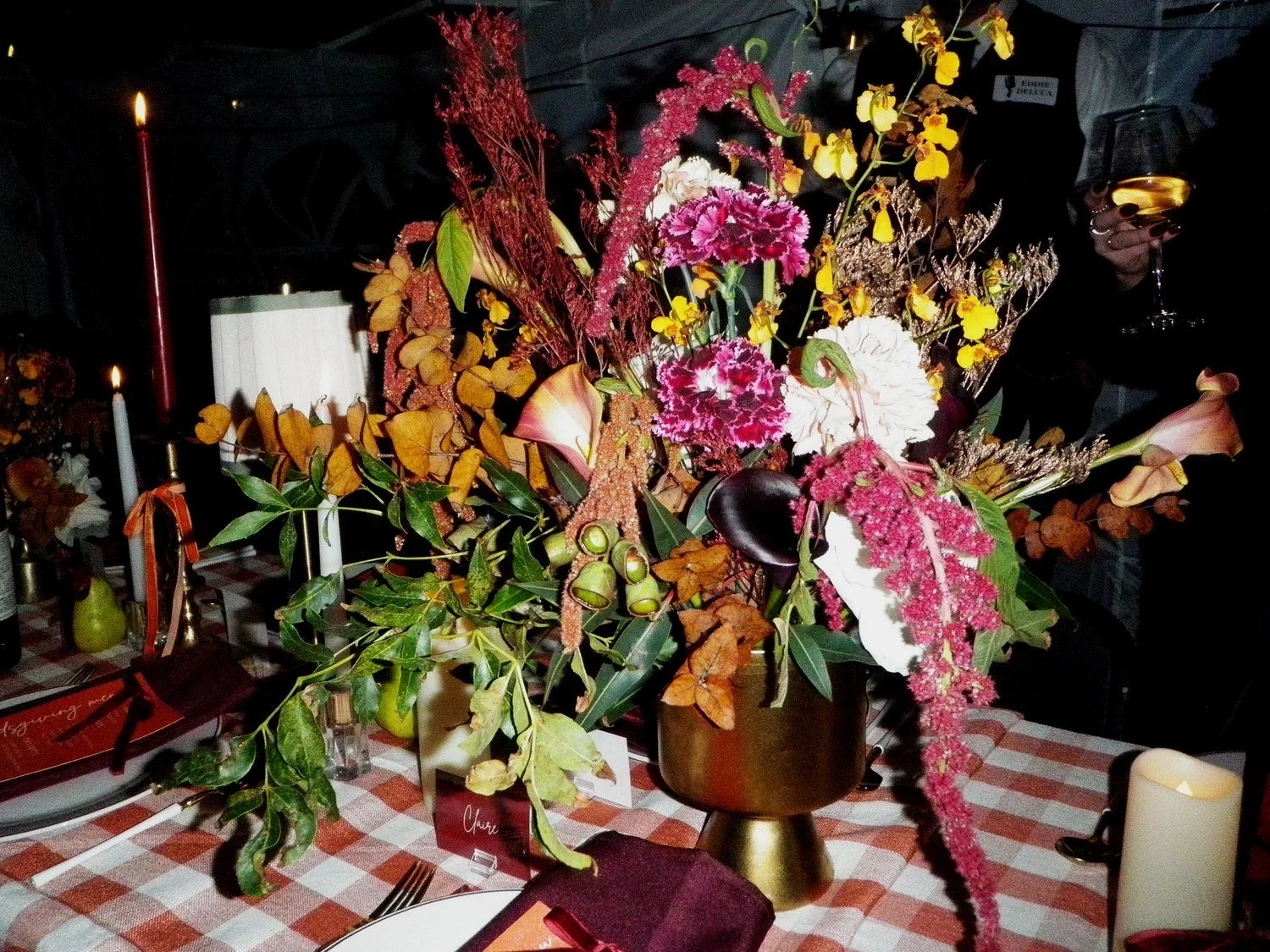 Colorful floral centerpiece with pink, white, and yellow flowers and green foliage on a checkered tablecloth at a dinner table with candles.