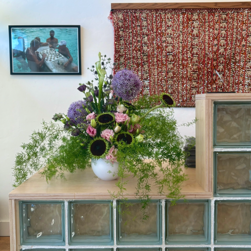 A floral arrangement with purple, pink, and green flowers on a wooden and glass block shelf, with a tapestry and a framed photo of people in a pool in the background.