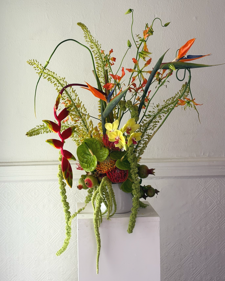 A tall floral arrangement in a white vase featuring various exotic flowers and green foliage against a neutral wall background.