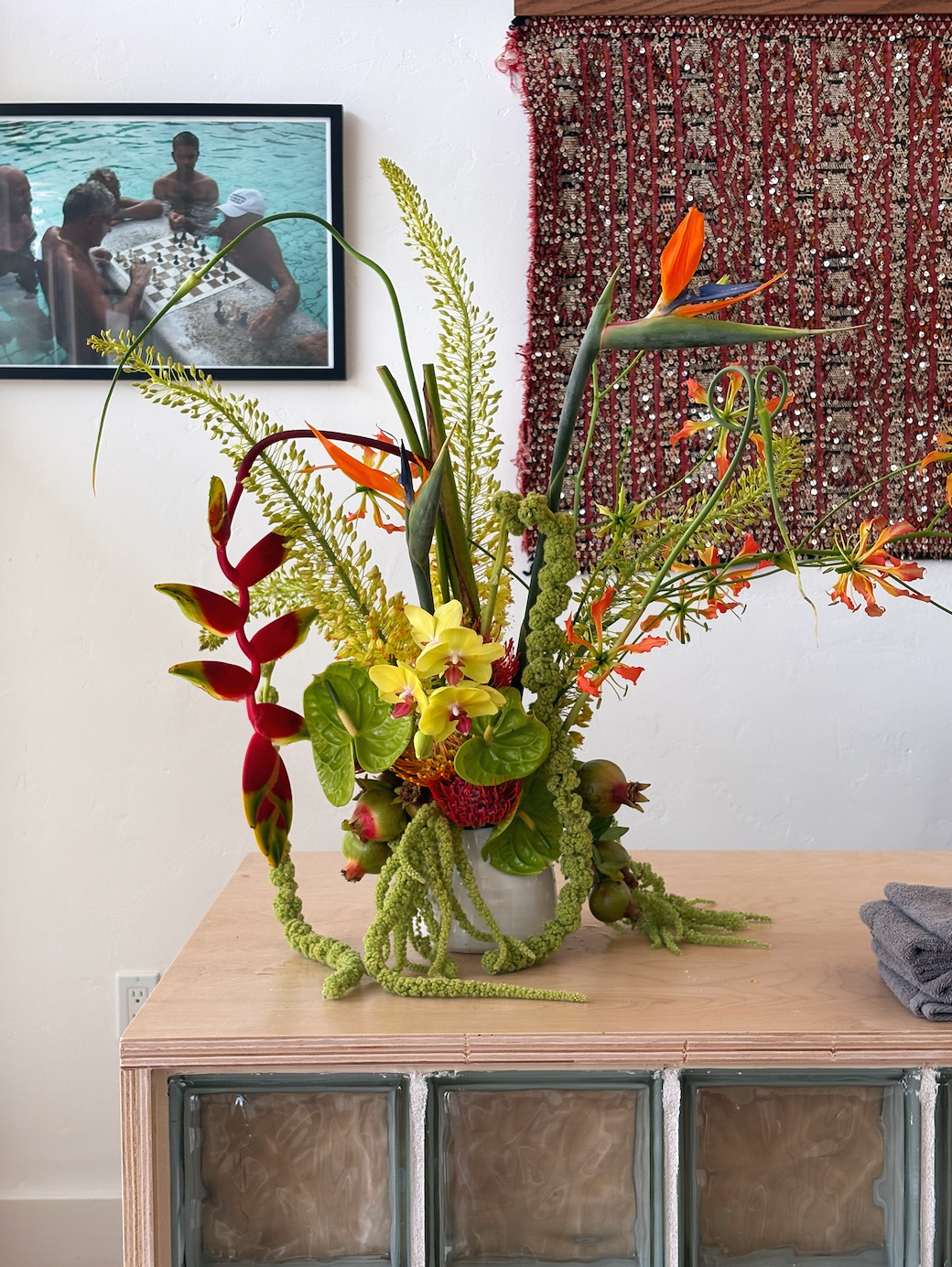 A colorful floral arrangement in a vase on a wooden table in front of a white wall with a framed photo of people playing chess in a swimming pool and a red textured wall hanging.