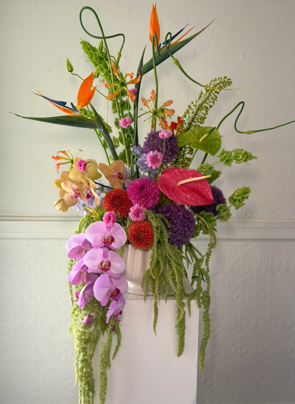 A colorful floral arrangement with orchids, anthuriums, dahlias, and other vibrant flowers in a white vase on a stand against a plain wall.